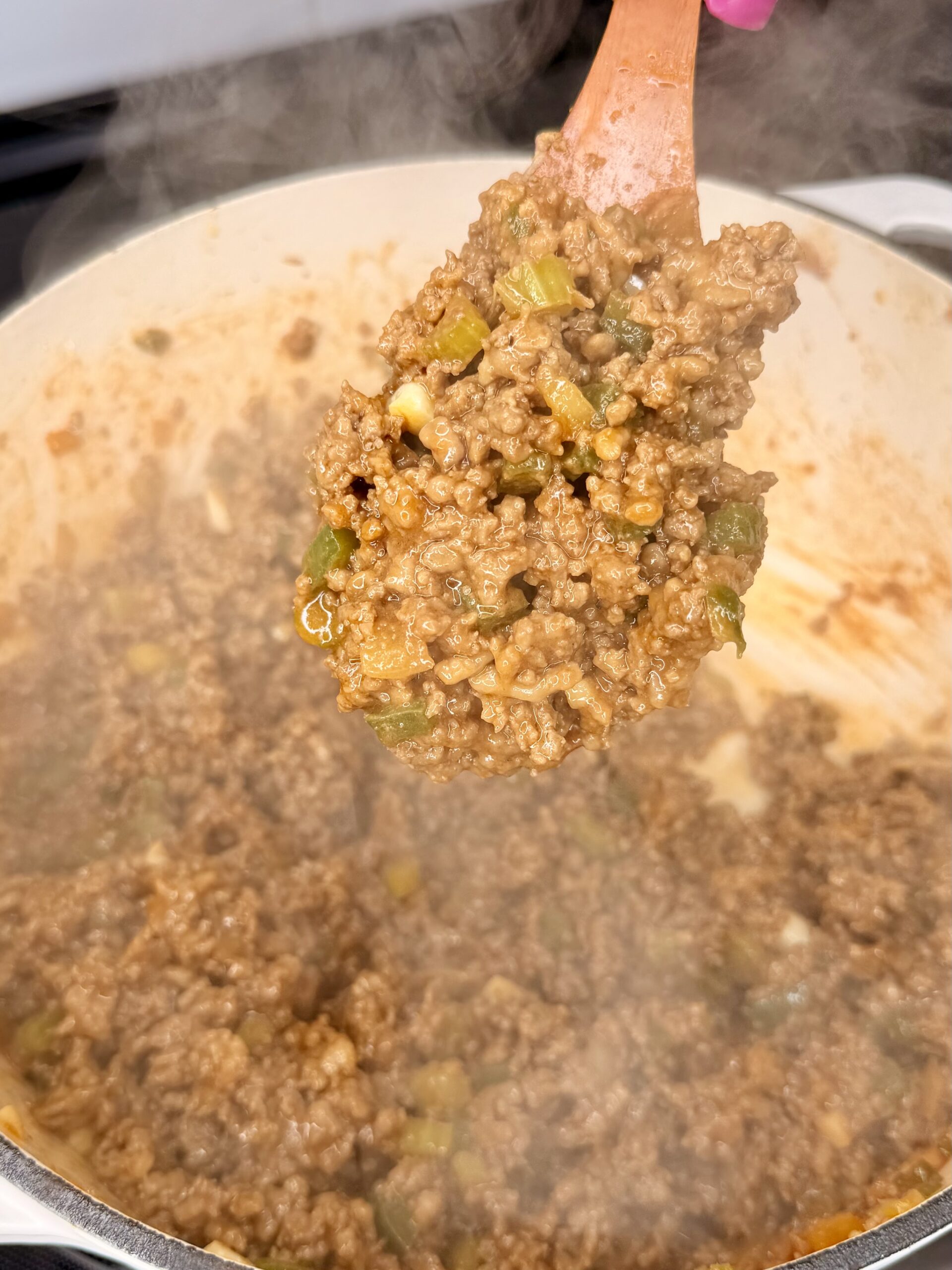 A wooden spoon lifts a serving of cooked ground beef mixed with diced green peppers and onions from a steamy pot. The mixture appears saucy and seasoned.