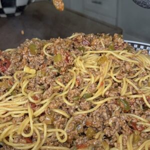 A close-up of cooked spaghetti mixed with ground beef, diced vegetables, and sauce being poured into a bowl from a pot. The background features a checkered vase and kitchen surfaces.