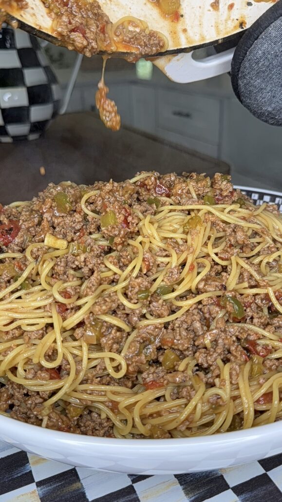 A close-up of cooked spaghetti mixed with ground beef, diced vegetables, and sauce being poured into a bowl from a pot. The background features a checkered vase and kitchen surfaces.