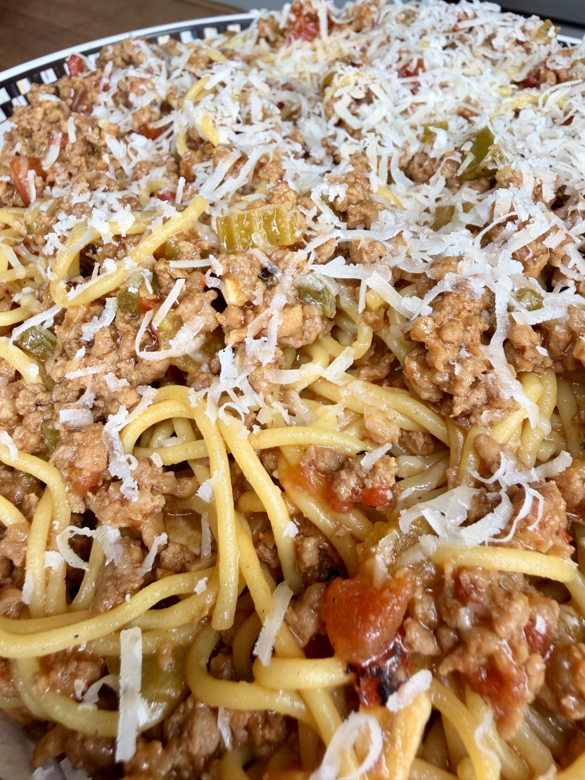 Close-up of cooked spaghetti topped with ground meat, tomato sauce, chopped vegetables, and grated cheese, served in a shallow dish.