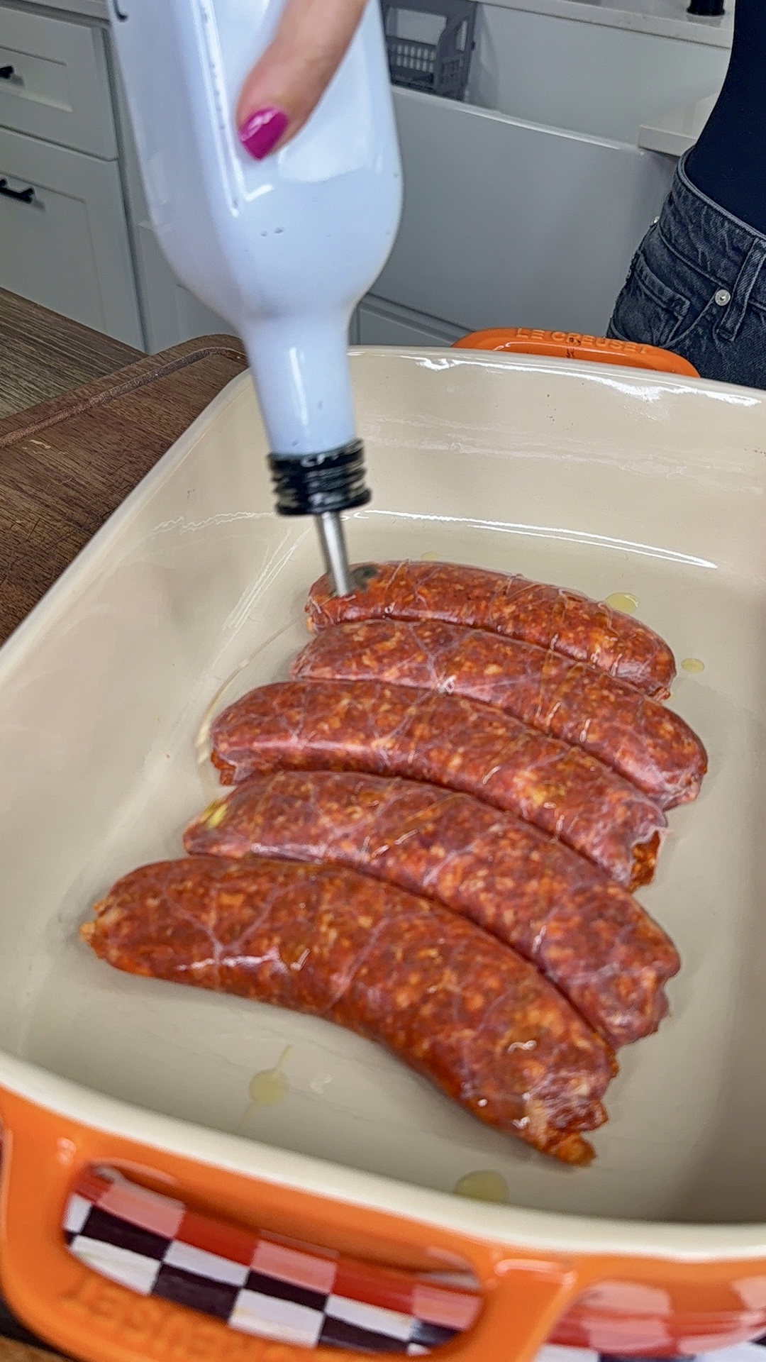 A hand pours olive oil from a white bottle over four raw sausages arranged in an orange baking dish on a kitchen counter.