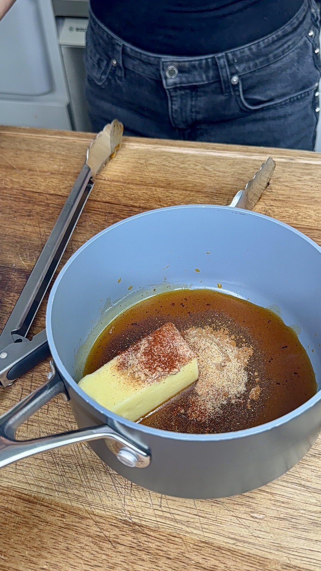 A saucepan on a wooden countertop contains a stick of butter, brown liquid, and various seasonings. Metal tongs rest nearby, and a person in dark jeans stands behind the counter.