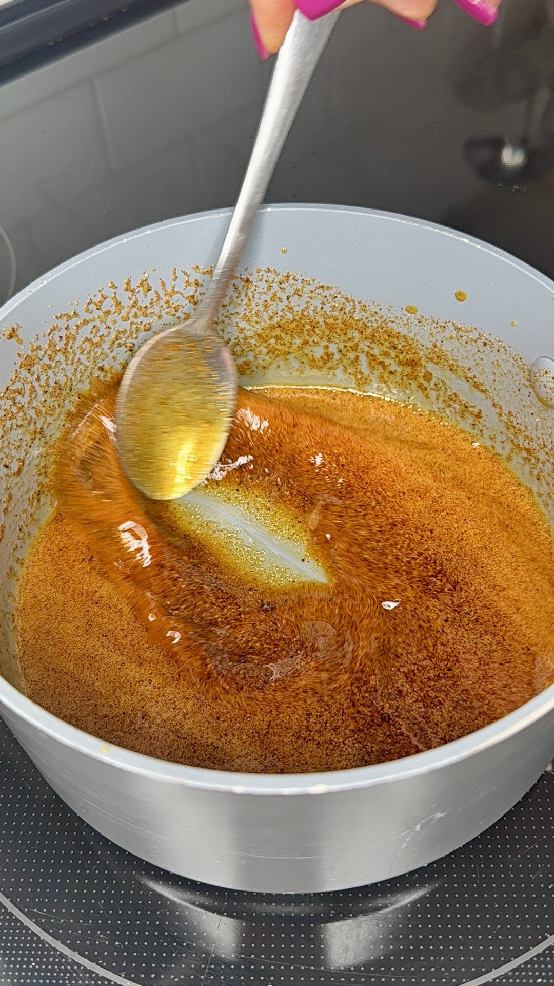 A hand stirs a pot of golden-brown caramelizing sugar with a metal spoon on a stovetop. The mixture is smooth and glossy, and the edges of the pot are coated with crystallized sugar.