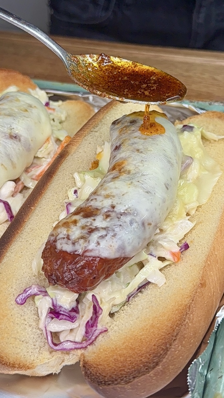 A close-up of a sausage covered in melted cheese on a bun with coleslaw, as a spoon drizzles reddish-brown sauce over the top.