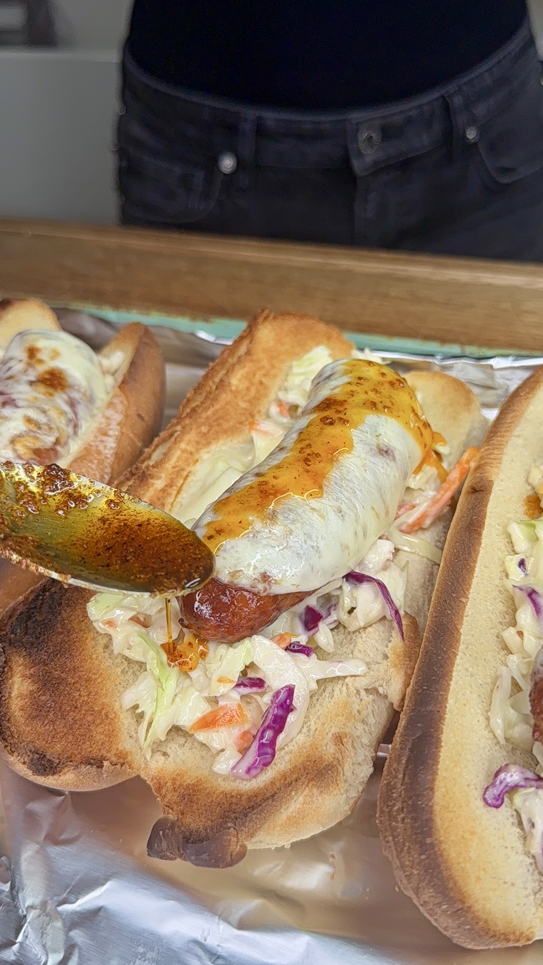 A sausage topped with melted cheese and coleslaw in a toasted bun is being drizzled with sauce from a spoon. Two similar sausage sandwiches are visible nearby on a foil-lined tray.