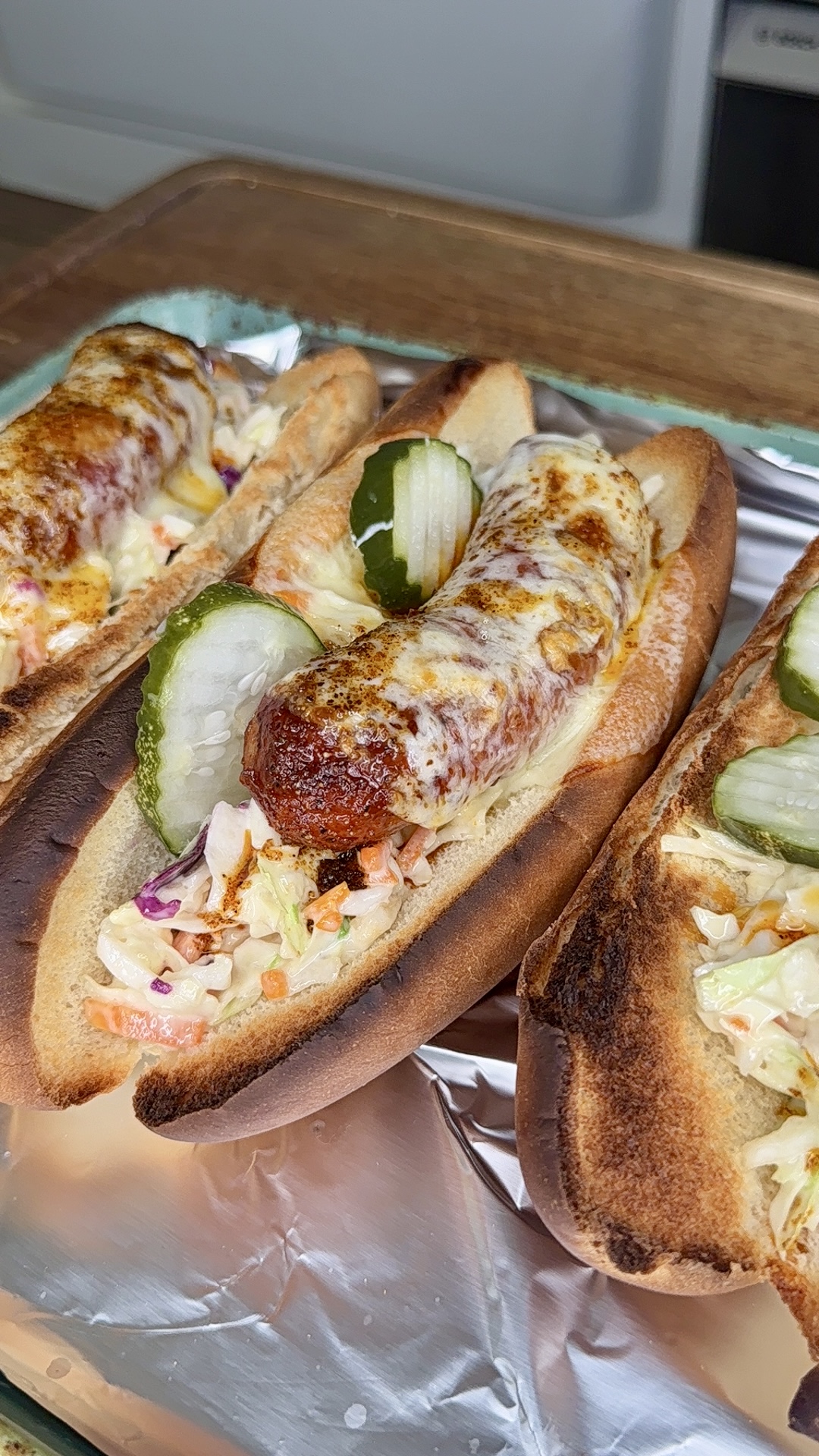 Three toasted hot dog buns filled with sausages topped with melted cheese, coleslaw, and pickle slices, set on a foil-lined tray.
