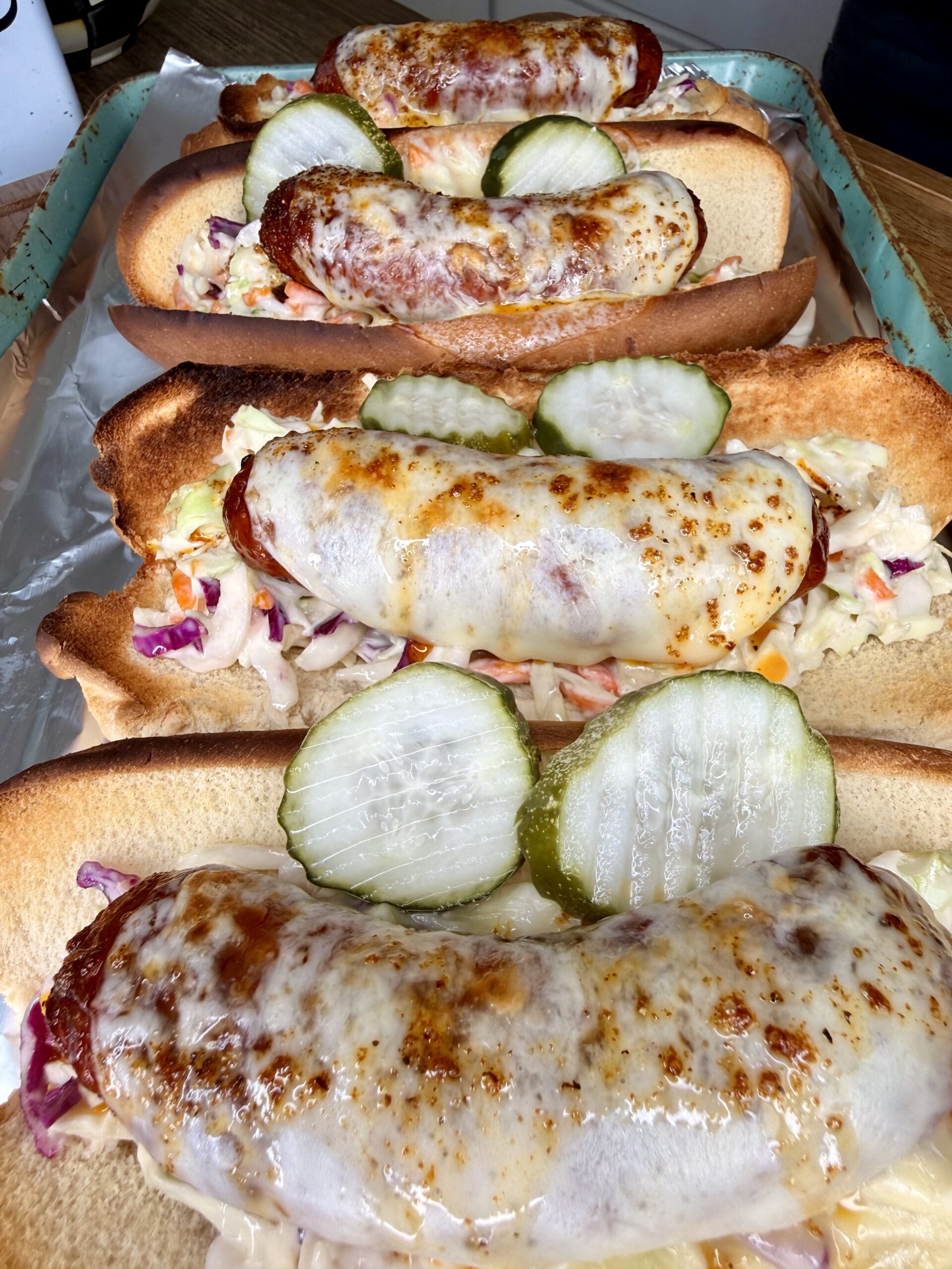 Four sausage hoagies topped with melted cheese, coleslaw, and pickle slices are arranged in a baking tray lined with foil.