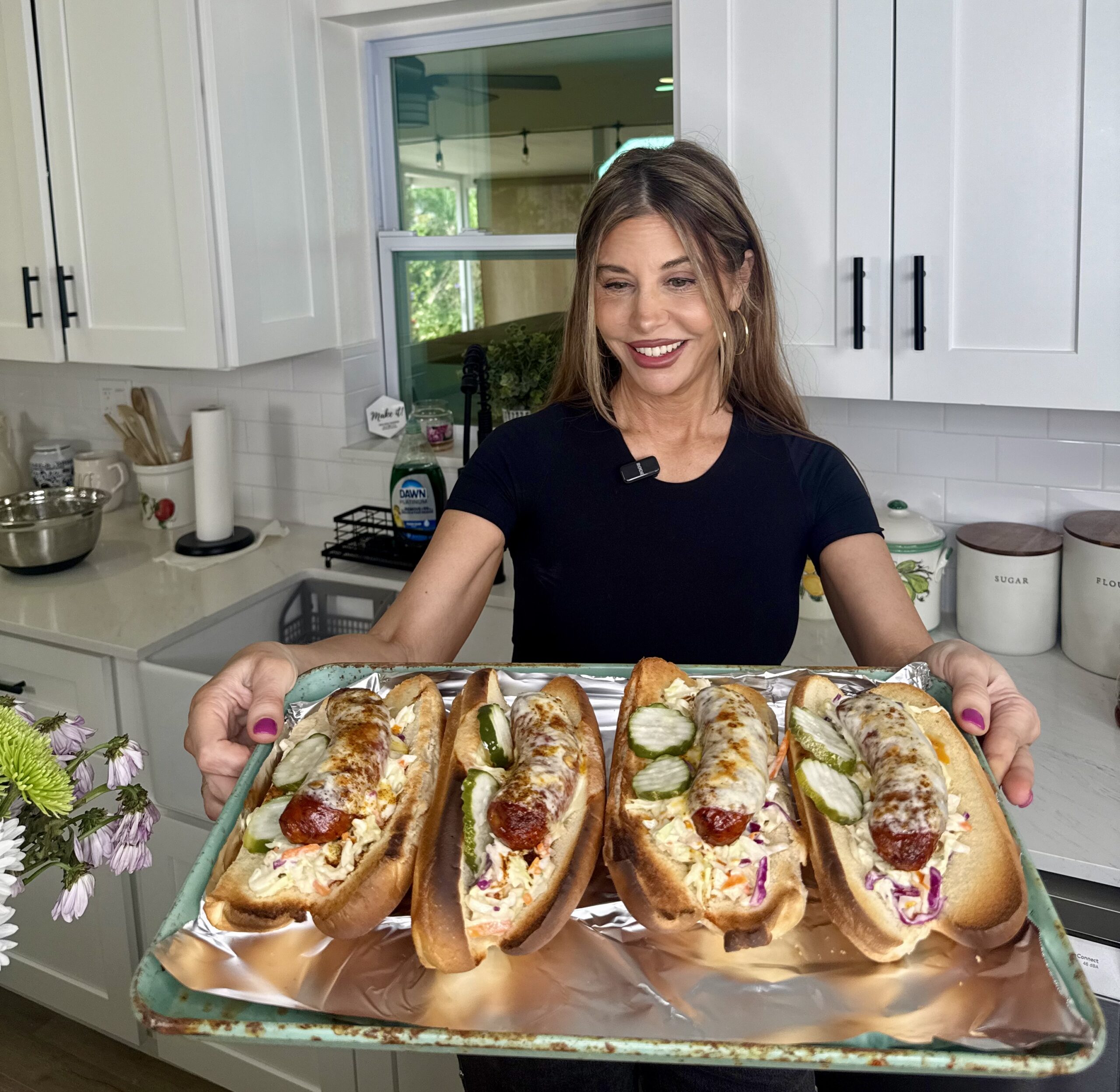 A smiling woman in a kitchen holds a tray with three large hot dogs topped with pickles, slaw, and sausages. The kitchen has white cabinets, a window, and various utensils in the background.