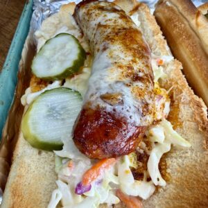 A toasted sandwich bun filled with a grilled sausage topped with melted cheese, sliced pickles, and creamy coleslaw, served on foil.
