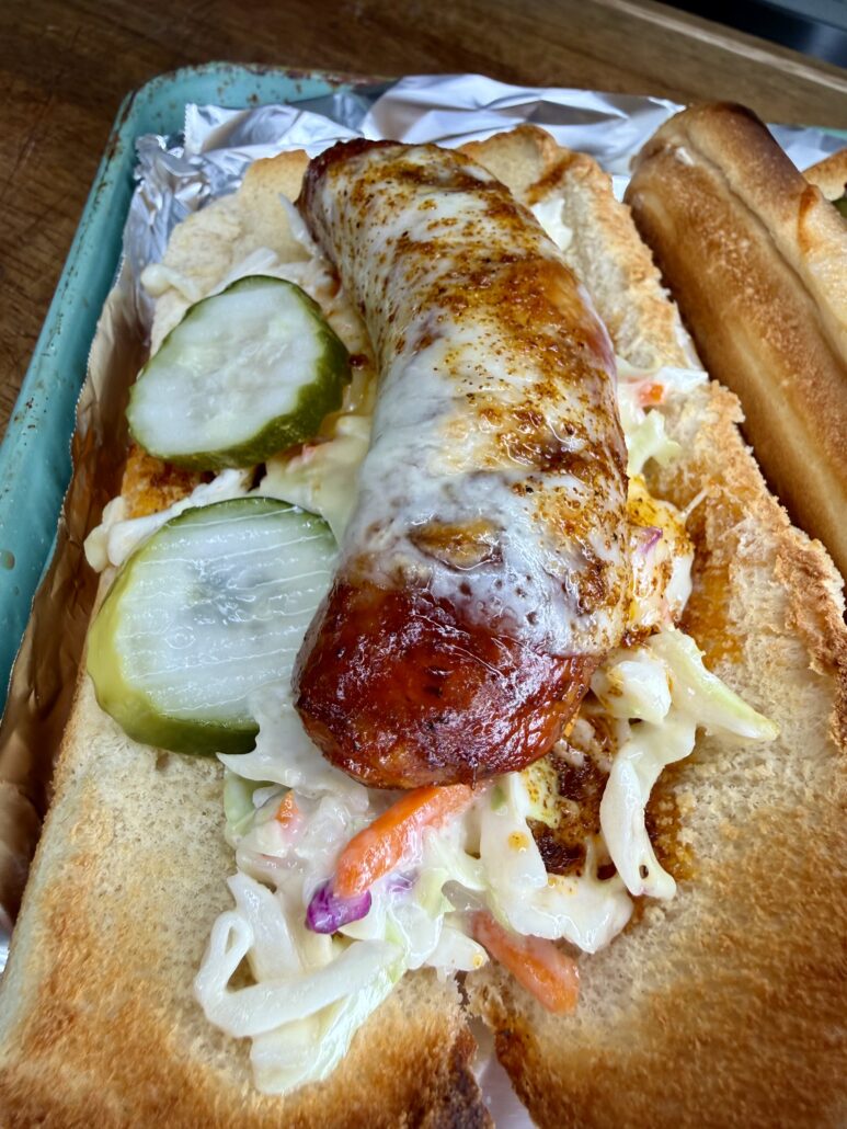 A toasted sandwich bun filled with a grilled sausage topped with melted cheese, sliced pickles, and creamy coleslaw, served on foil.