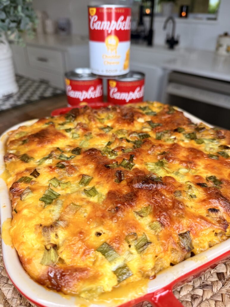 A Cheddar Make-Ahead Breakfast casserole topped with melted cheese and green onions sits in a red dish on the kitchen counter. Behind it are three stacked cans of Campbell’s Cheddar Cheese soup.