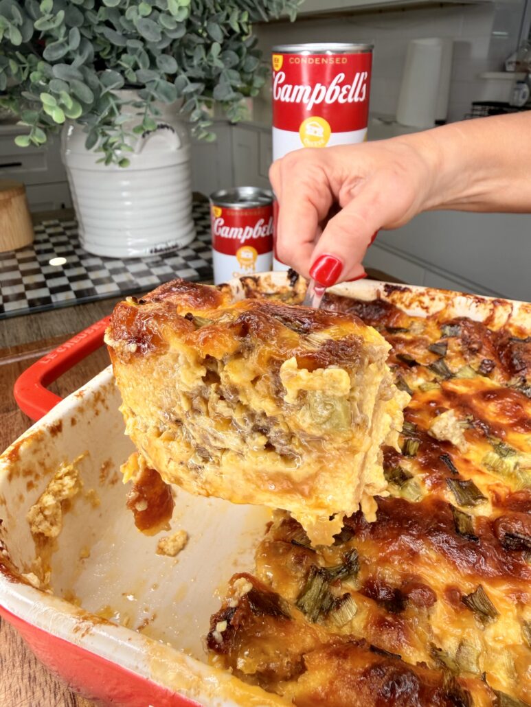 A hand with red nail polish lifts a large serving of Cheddar Make-Ahead Breakfast casserole from a red dish, with Campbell's soup cans and kitchen items in the background.