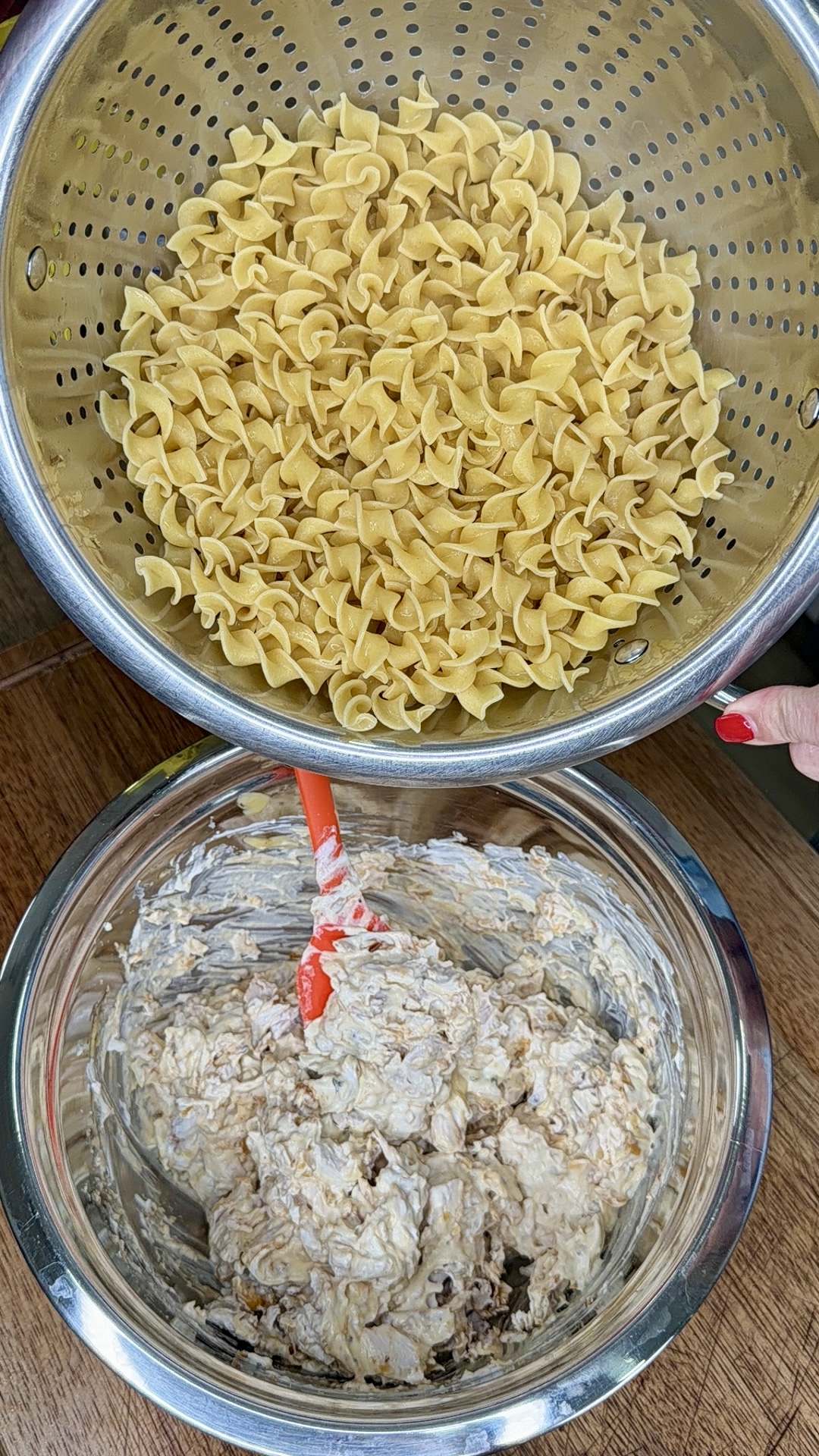 A metal colander filled with cooked egg noodles sits above a glass bowl containing a mixture being stirred with a red spatula. A hand with red nail polish is holding the colander. Both are on a wooden surface.