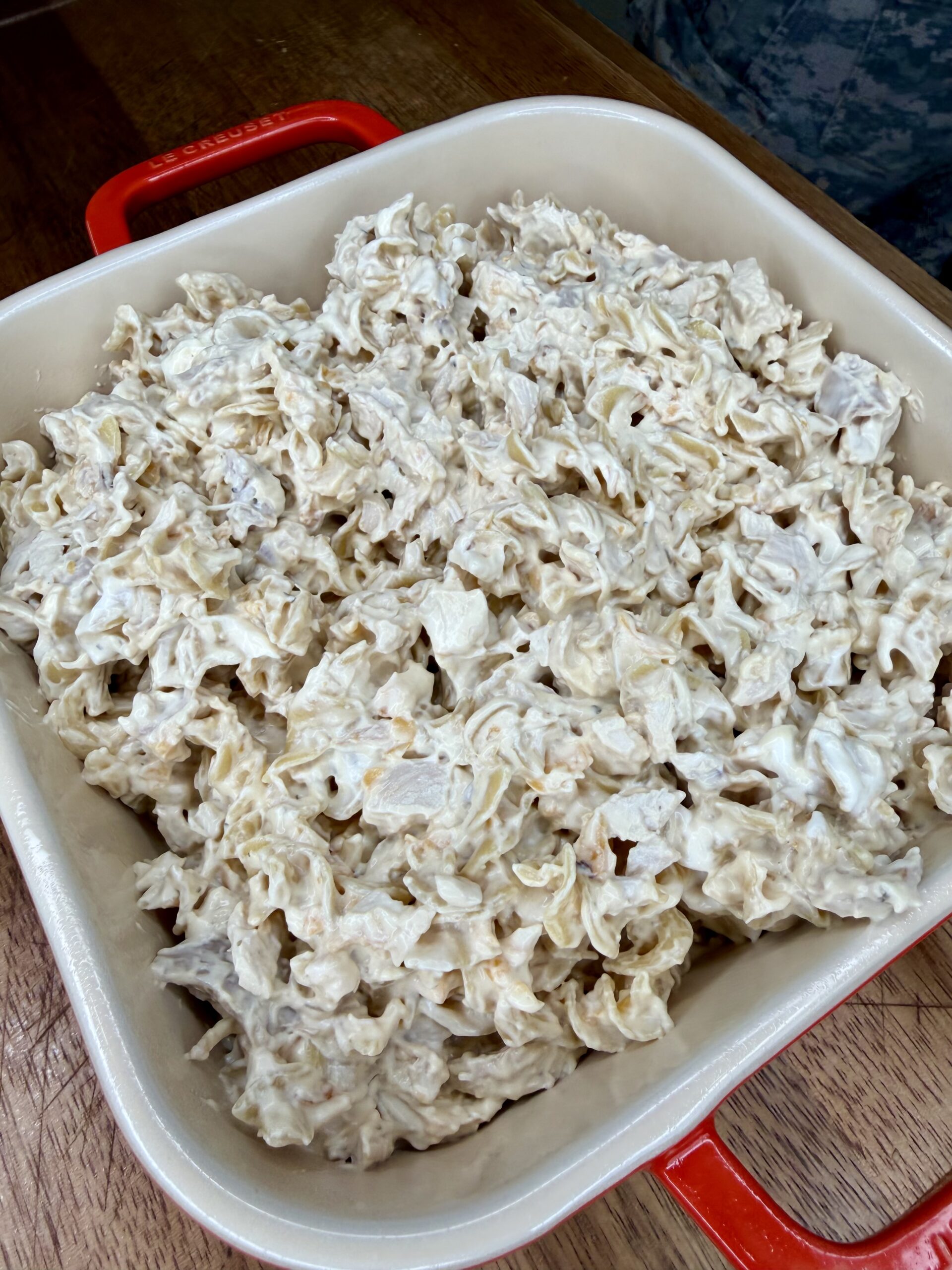 A square baking dish with red handles filled with a creamy pasta casserole, featuring curly noodles mixed with a white sauce, sitting on a wooden surface.