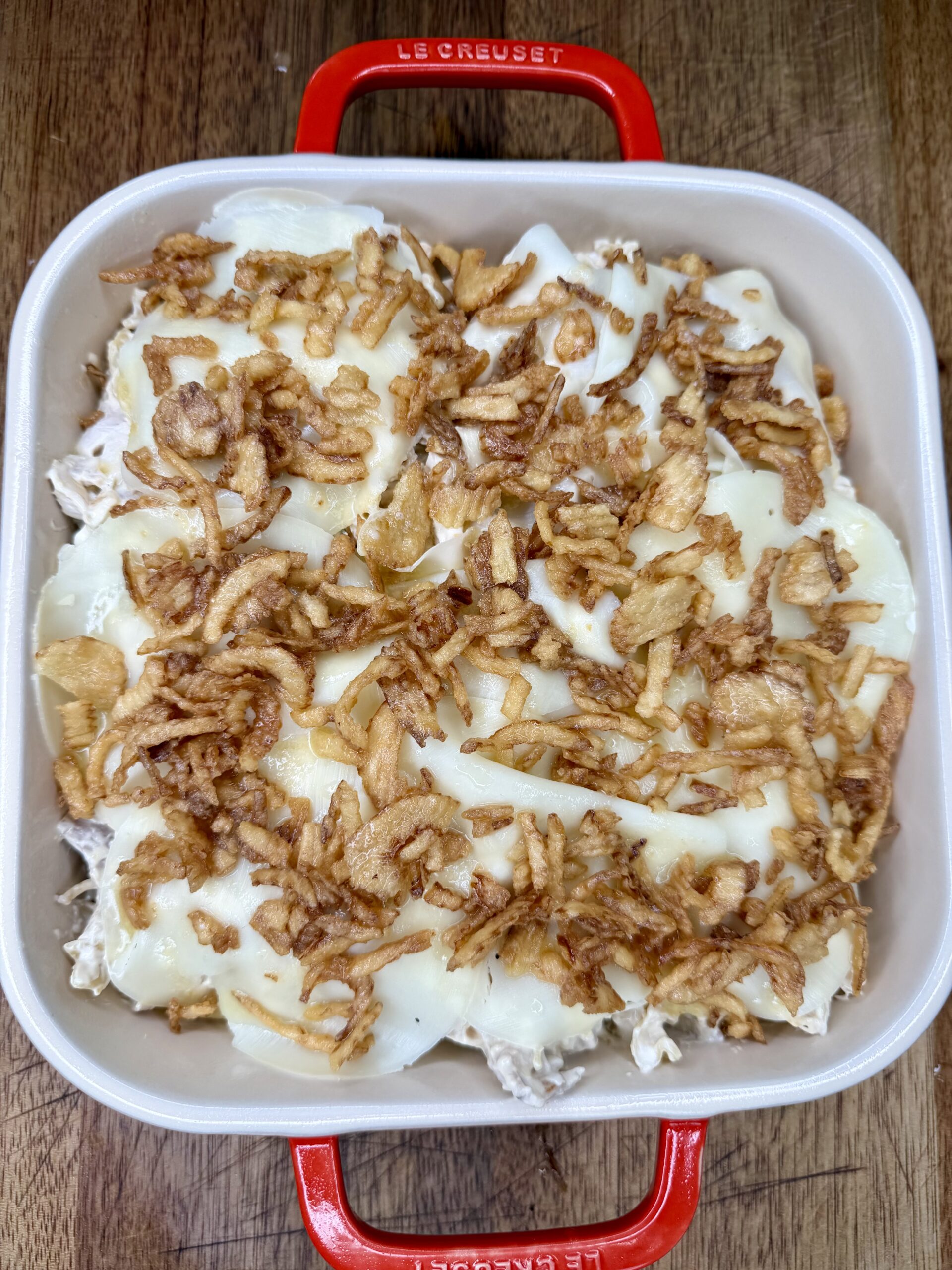 A square red baking dish filled with a casserole topped with melted cheese and crispy fried onions, set on a wooden surface.