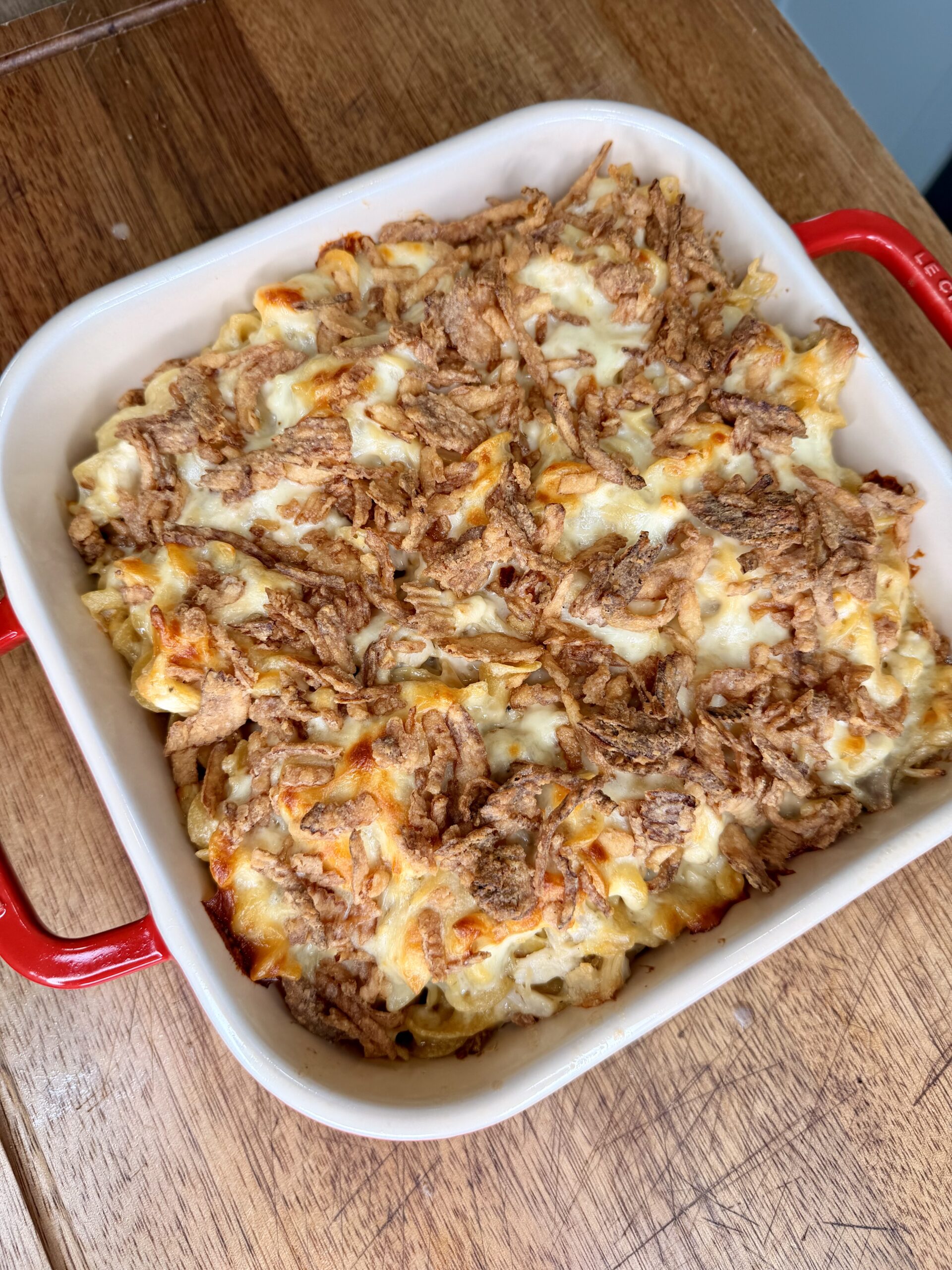 A baked casserole in a square red-handled dish, topped with crispy fried onions, sitting on a wooden surface. The casserole appears golden brown with a textured, crunchy topping.