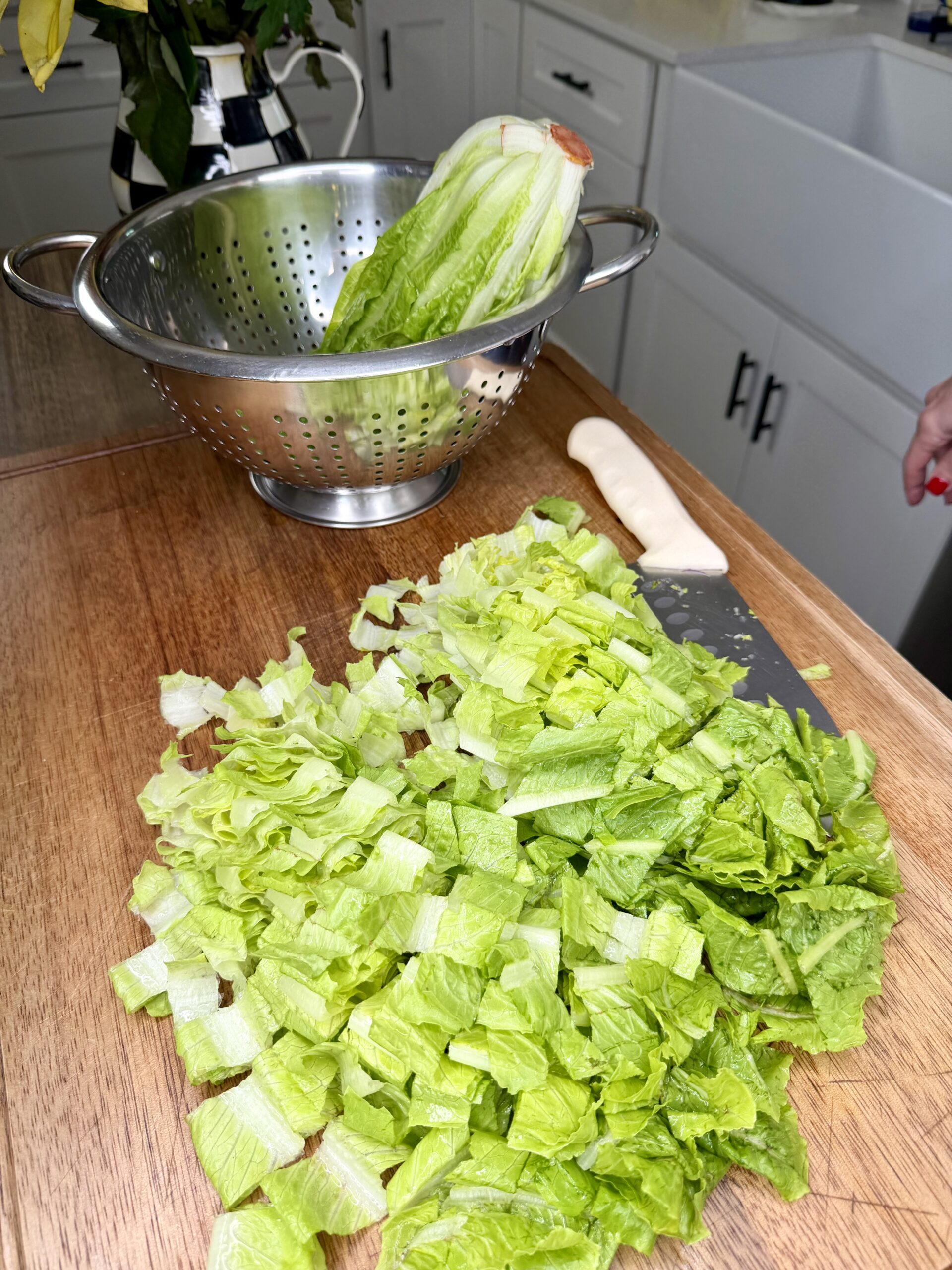 Chopped romaine lettuce is spread on a wooden countertop beside a large knife. A metal colander with a head of unchopped romaine sits behind the cutting board in a bright kitchen.
