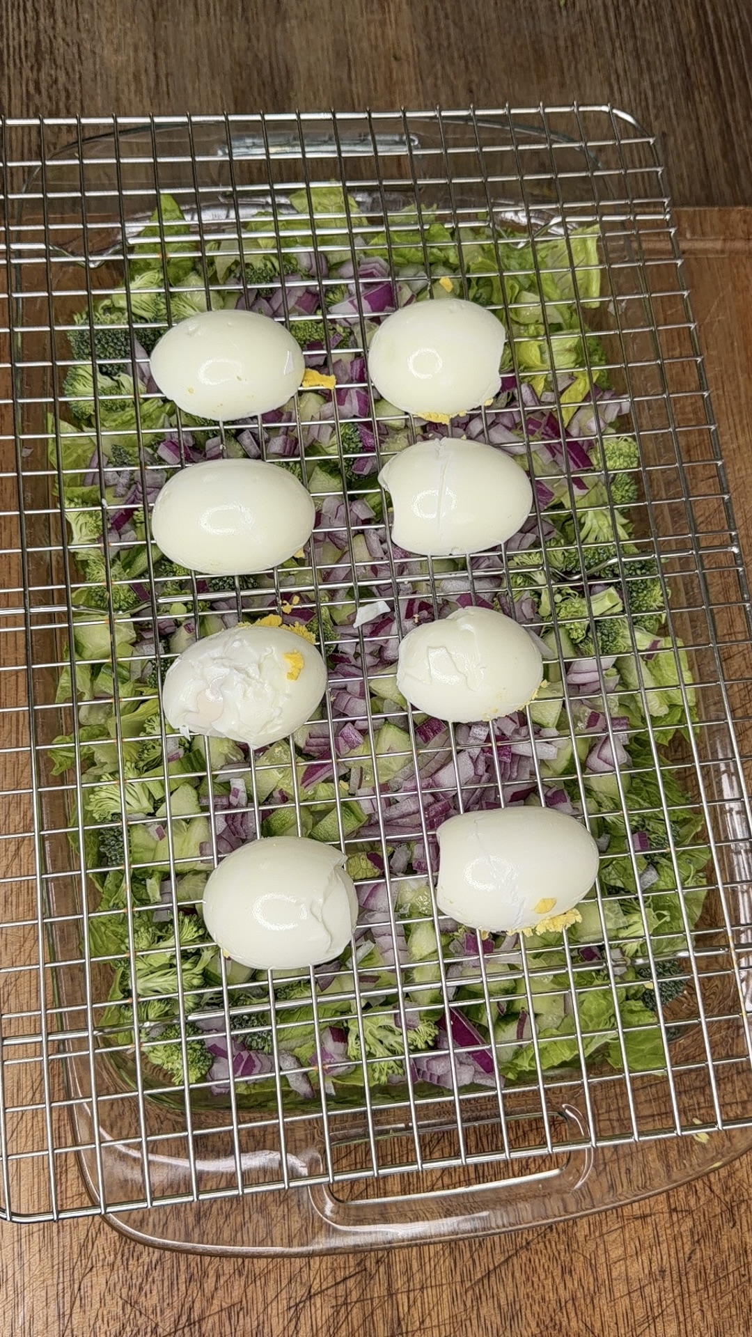 Eight peeled hard-boiled eggs rest on a metal cooling rack set over a tray filled with chopped lettuce, red onions, and broccoli on a wooden surface.
