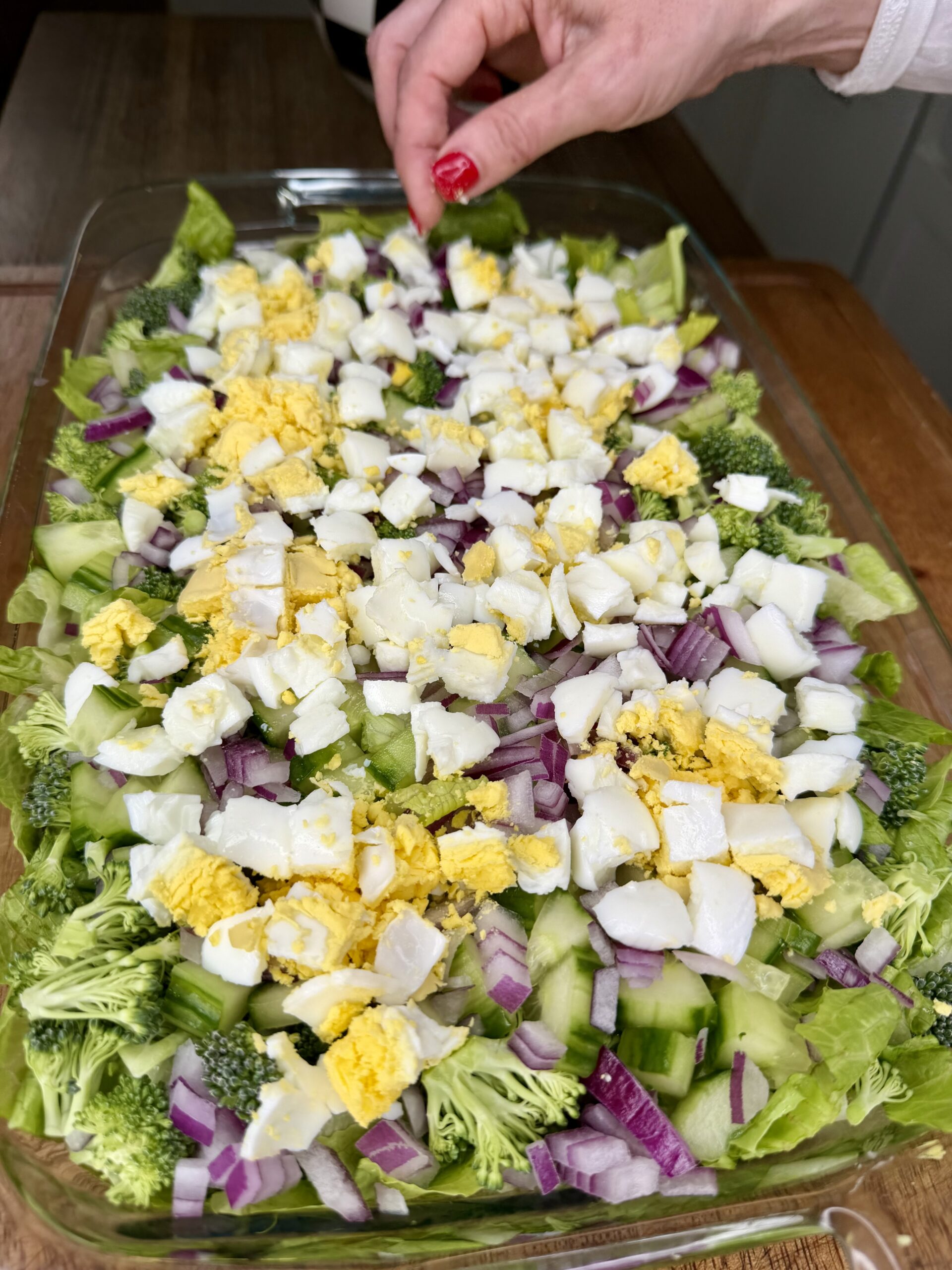 A hand sprinkles pieces over a glass dish filled with a layered salad of lettuce, chopped hard-boiled eggs, broccoli, cucumber, and red onion on a wooden table.