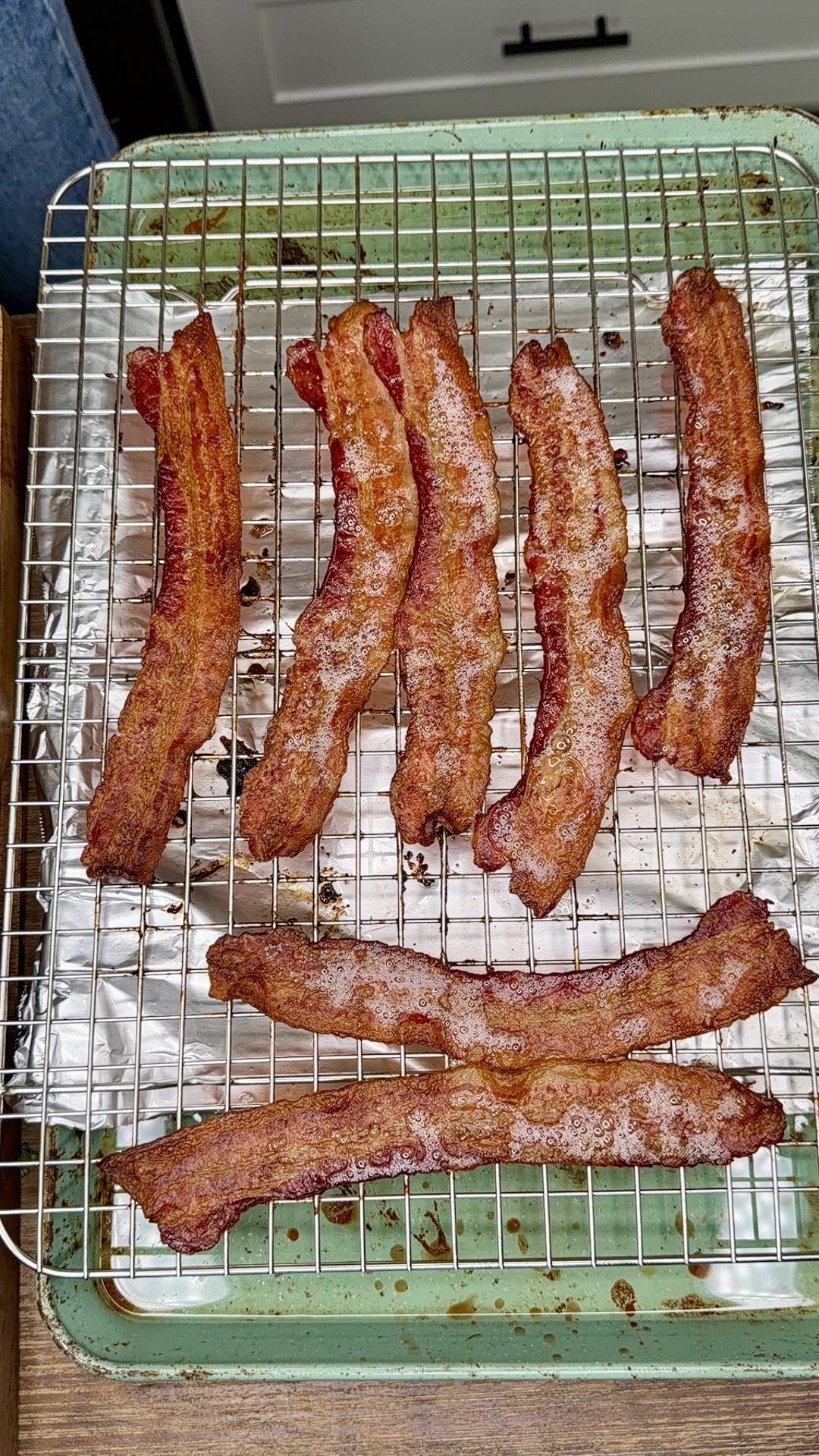 Six crispy strips of bacon cooling on a metal rack over a foil-lined green baking sheet. Four strips are arranged vertically at the top and two horizontally at the bottom.