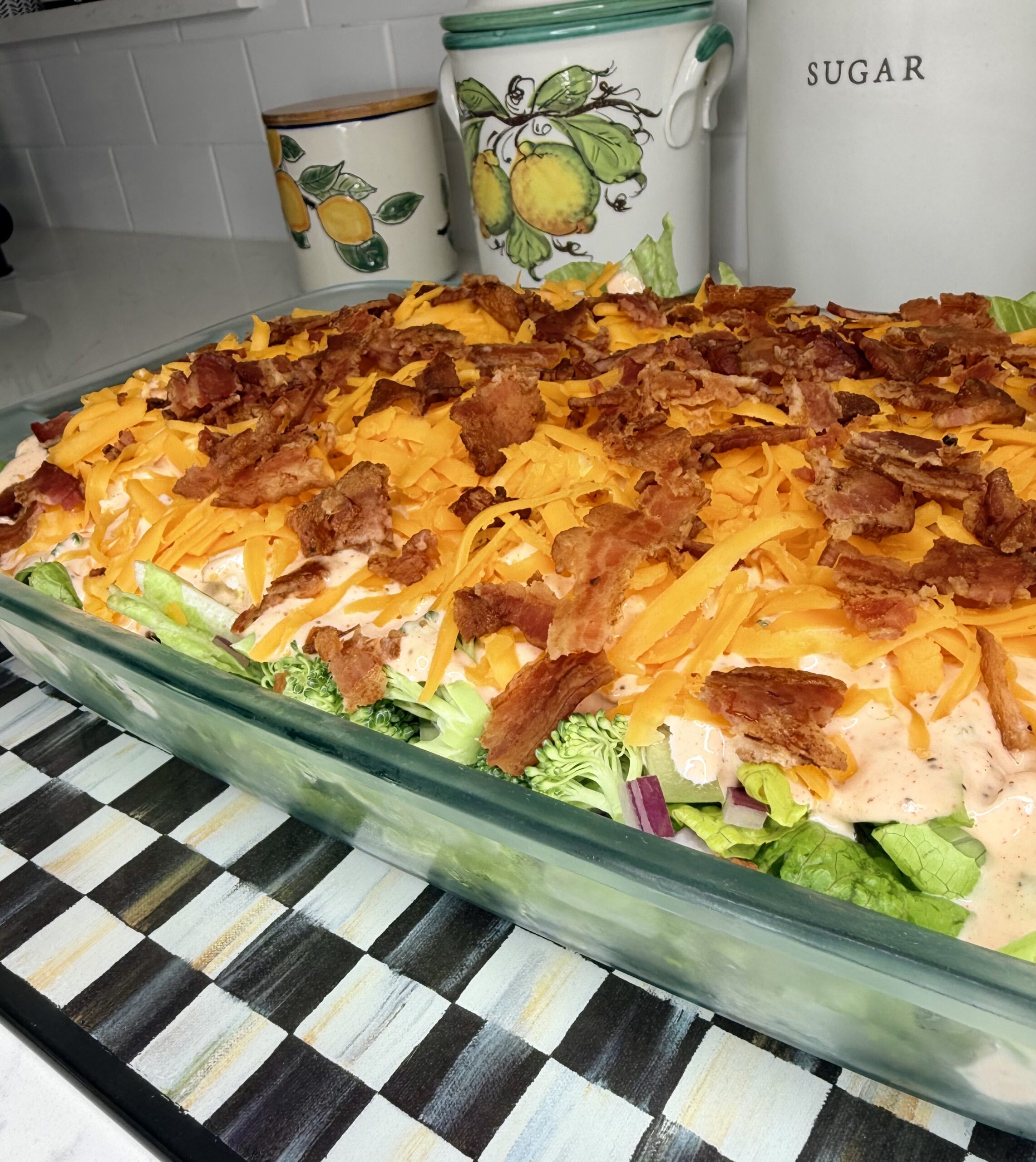 A glass baking dish filled with layered salad topped with shredded cheddar cheese and crispy bacon pieces, set on a striped mat in a kitchen with decorative containers in the background.