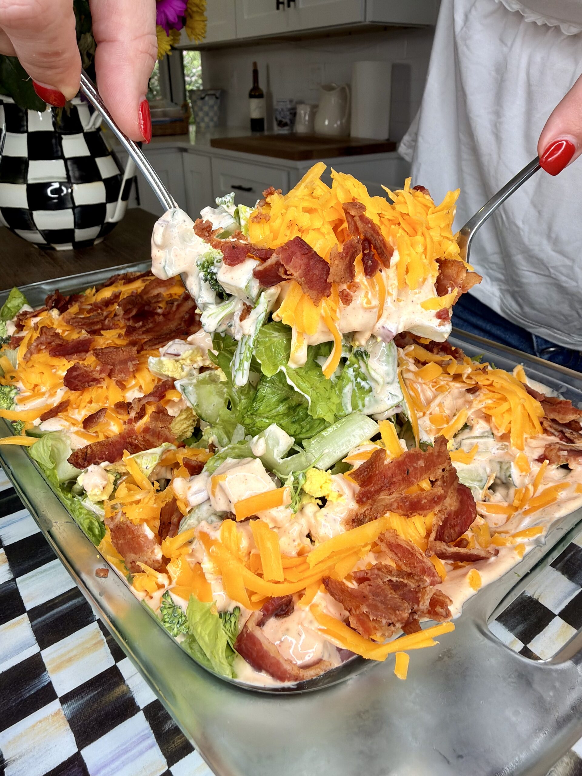 A person serves a portion of layered salad topped with shredded cheddar cheese and crispy bacon from a glass dish in a bright kitchen. The salad contains lettuce and creamy dressing.