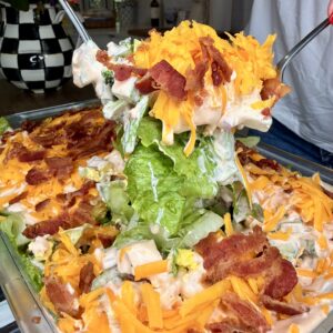 A person uses two utensils to serve a large portion of layered salad topped with shredded cheddar cheese, crispy bacon, and creamy dressing from a glass dish. Fresh lettuce and other vegetables are visible underneath.