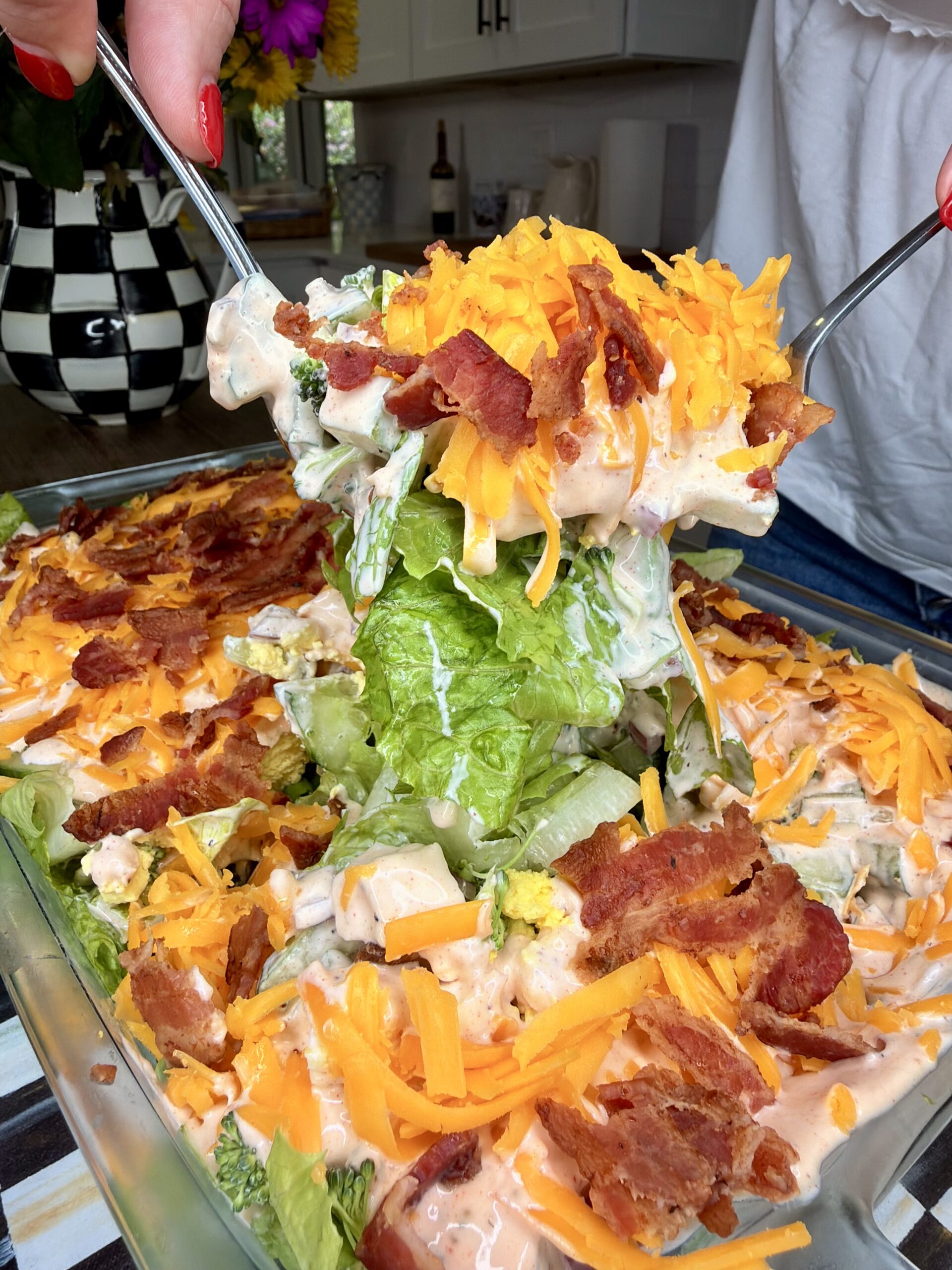 A person uses two utensils to serve a large portion of layered salad topped with shredded cheddar cheese, crispy bacon, and creamy dressing from a glass dish. Fresh lettuce and other vegetables are visible underneath.