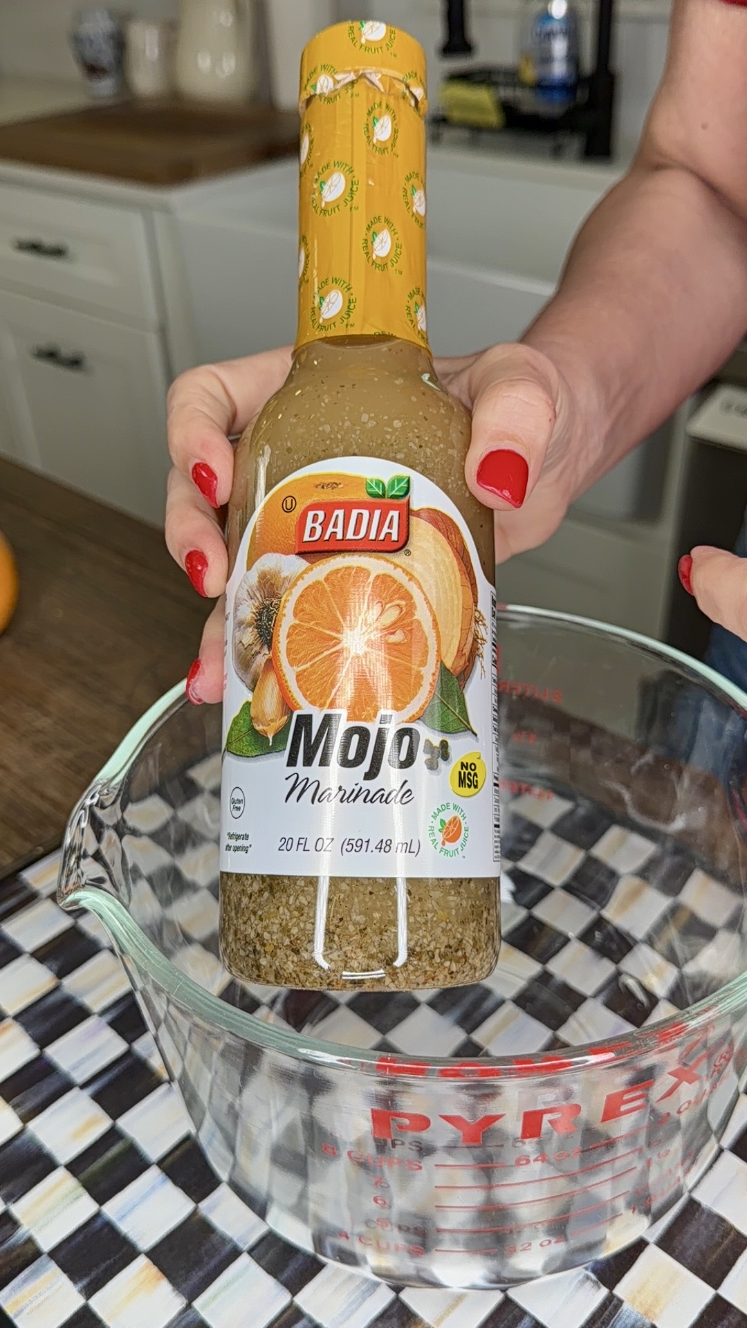 A person with red nail polish holds a bottle of Badia Mojo Marinade above a glass Pyrex measuring cup, ready to prep a delicious One-Pan Mojo Chicken & Potatoes meal on a checkered kitchen surface.