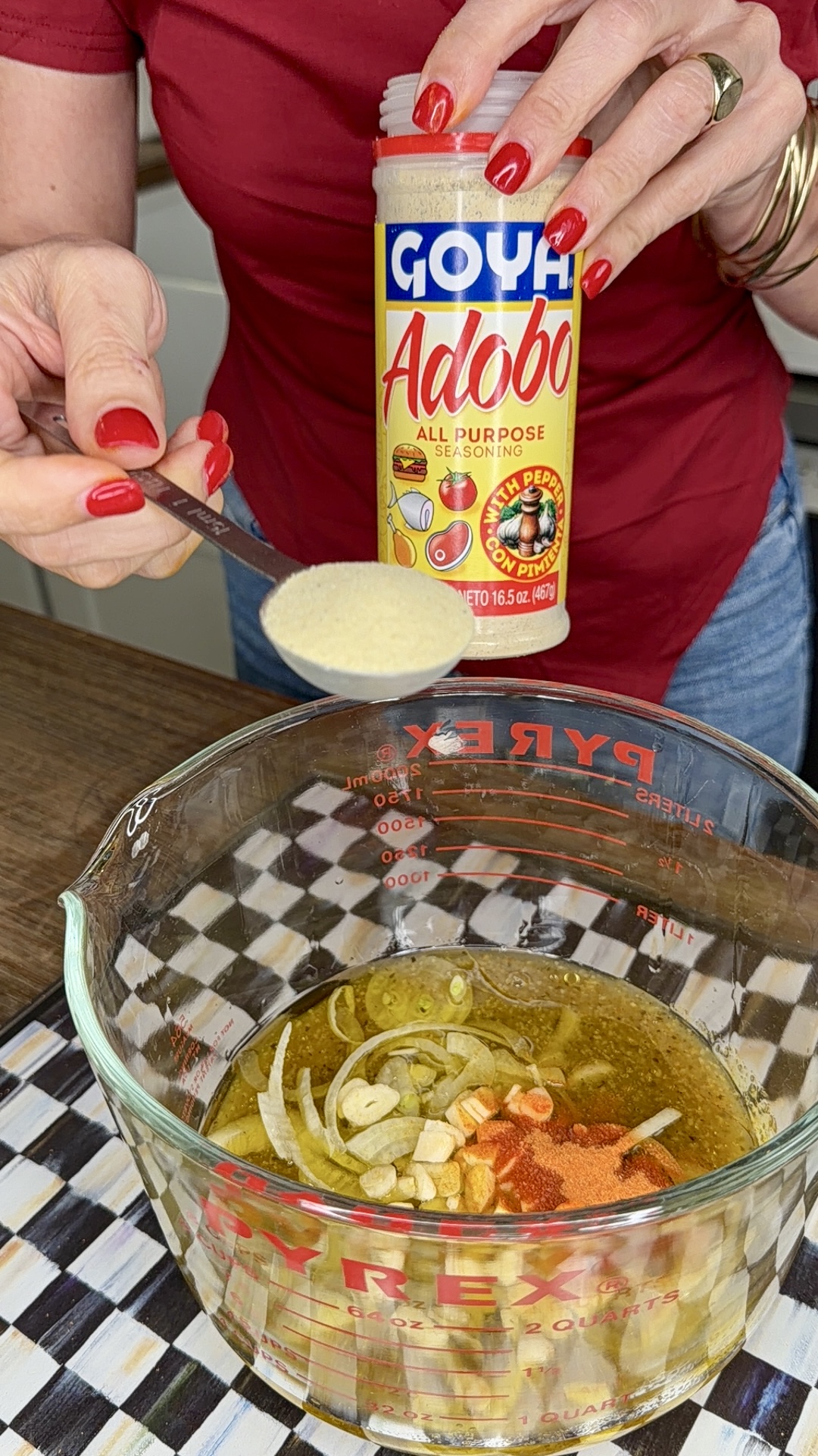 A person sprinkles Goya Adobo seasoning from a jar into a glass measuring cup filled with sliced onions, garlic, oil, and spices—prepping the flavorful marinade for One-Pan Mojo Chicken & Potatoes on a checkered surface.