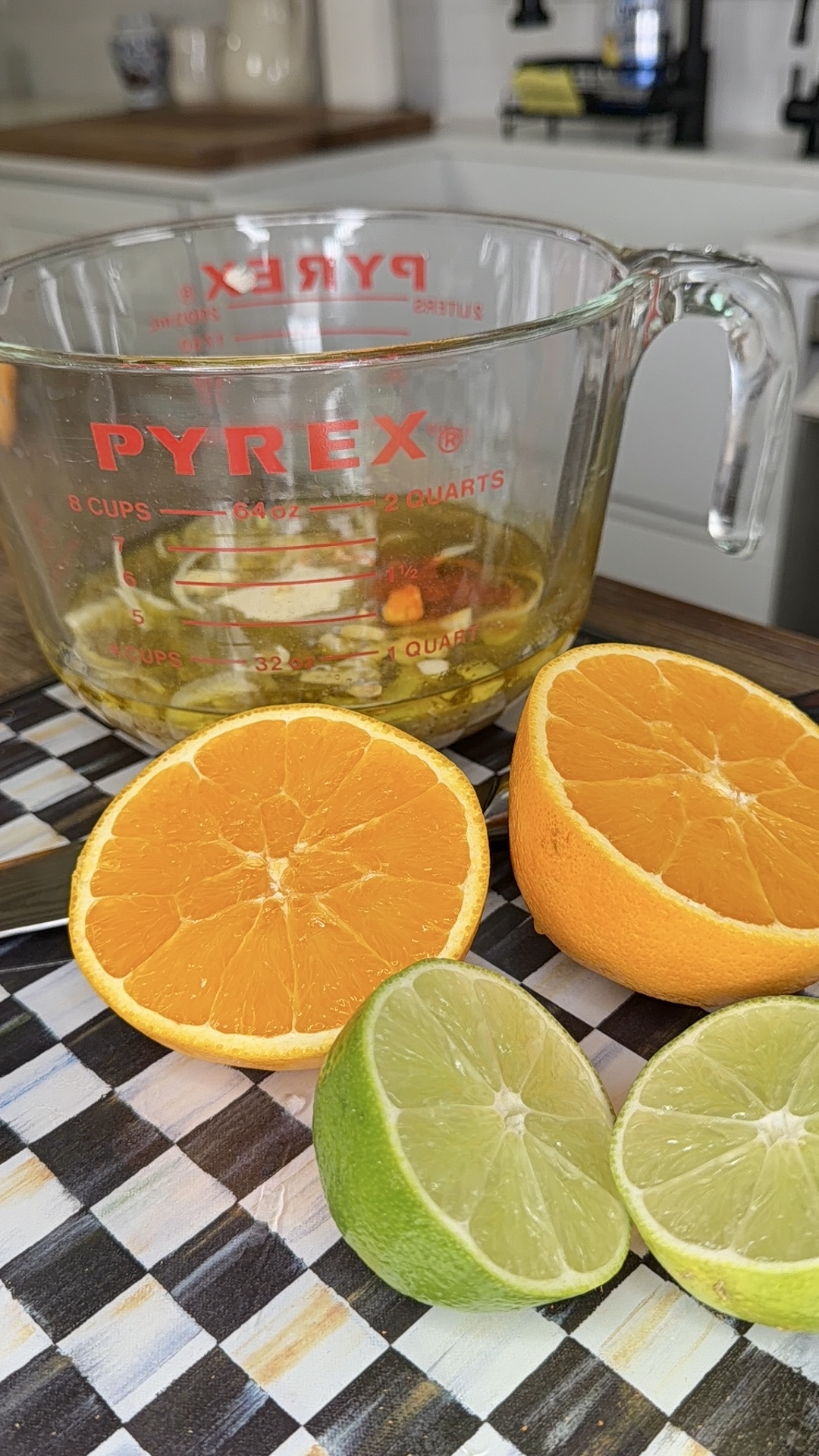 A glass Pyrex measuring cup with liquid inside sits on a counter behind halved lime and orange on a checkered cutting board—perfect for prepping citrus juice for One-Pan Mojo Chicken & Potatoes. A knife is nearby in a cozy kitchen setting.