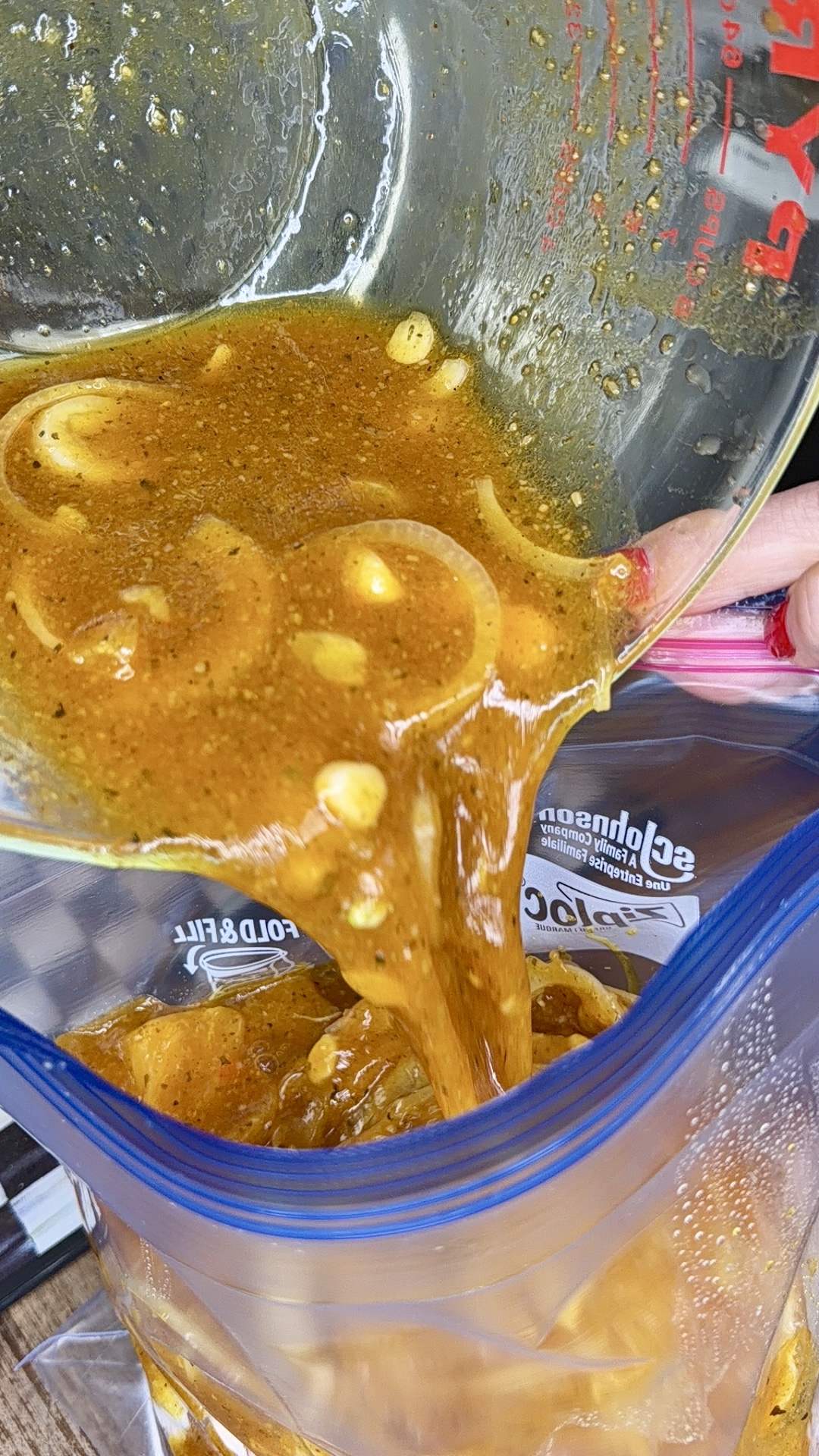 A hand pours a thick, orange-brown marinade with sliced onions from a glass measuring cup into a resealable plastic bag, ready to make One-Pan Mojo Chicken & Potatoes.