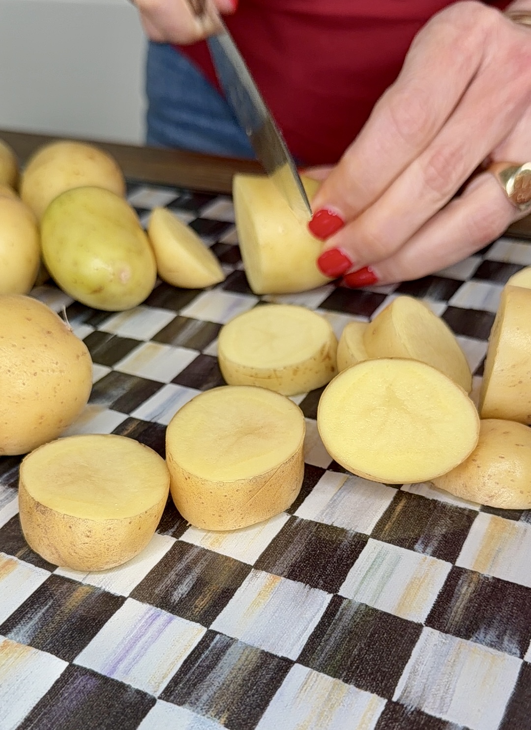 A person with red nail polish is slicing small yellow potatoes for a delicious One-Pan Mojo Chicken & Potatoes meal on a black and white checkered cutting board. Several whole and sliced potatoes are visible.