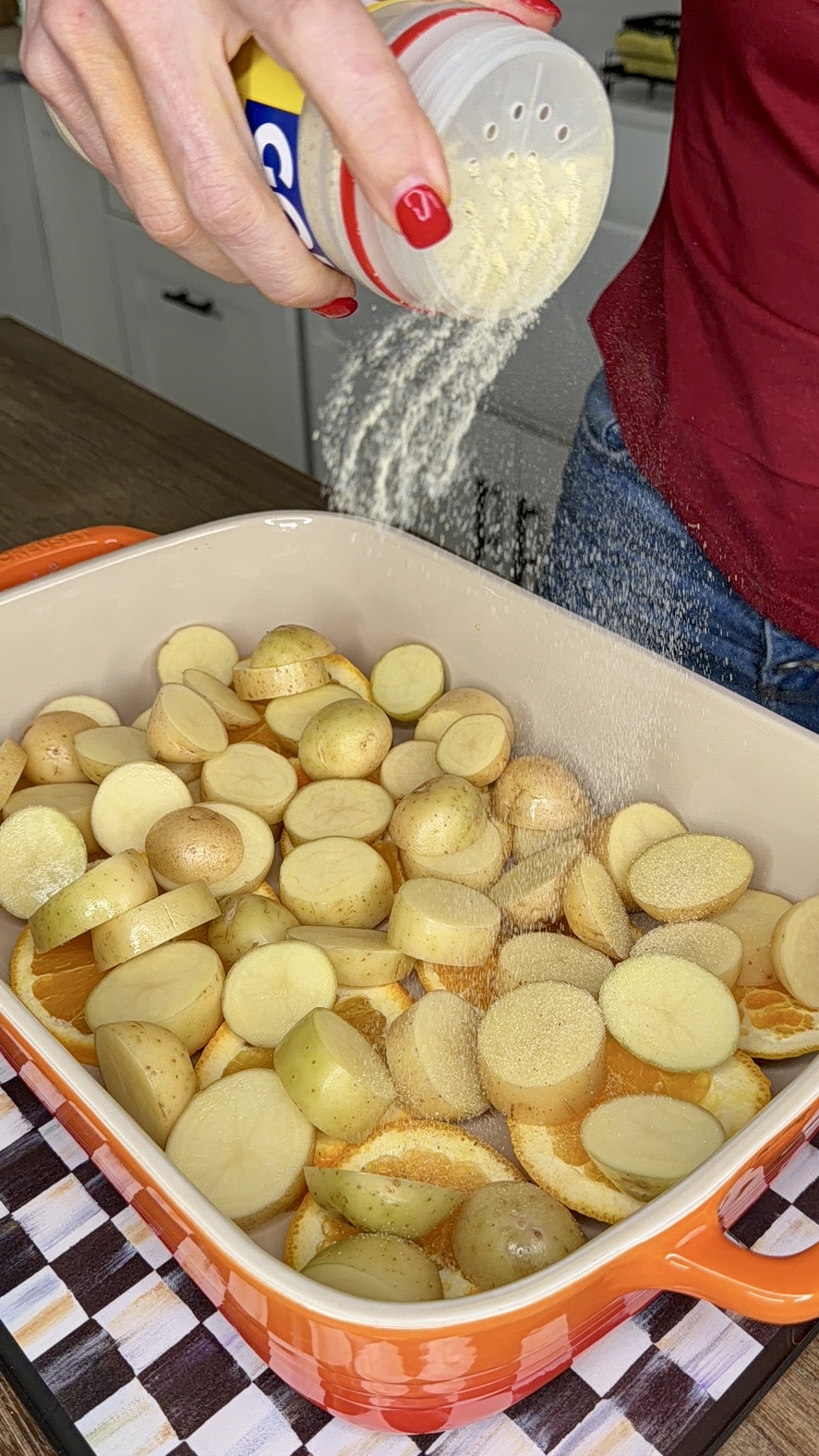 A person sprinkles seasoning from a container onto sliced potatoes in an orange baking dish, preparing them for cooking One-Pan Mojo Chicken & Potatoes. The dish sits on a checkered cloth on a countertop.