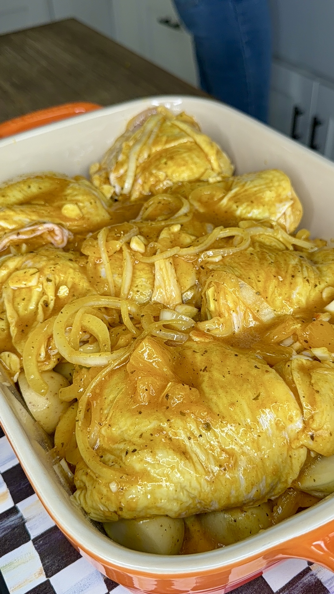 A baking dish filled with seasoned One-Pan Mojo Chicken & Potatoes, topped with sliced onions and sauce, ready to be cooked. The dish sits on a countertop, with part of a kitchen and a person’s legs in the background.