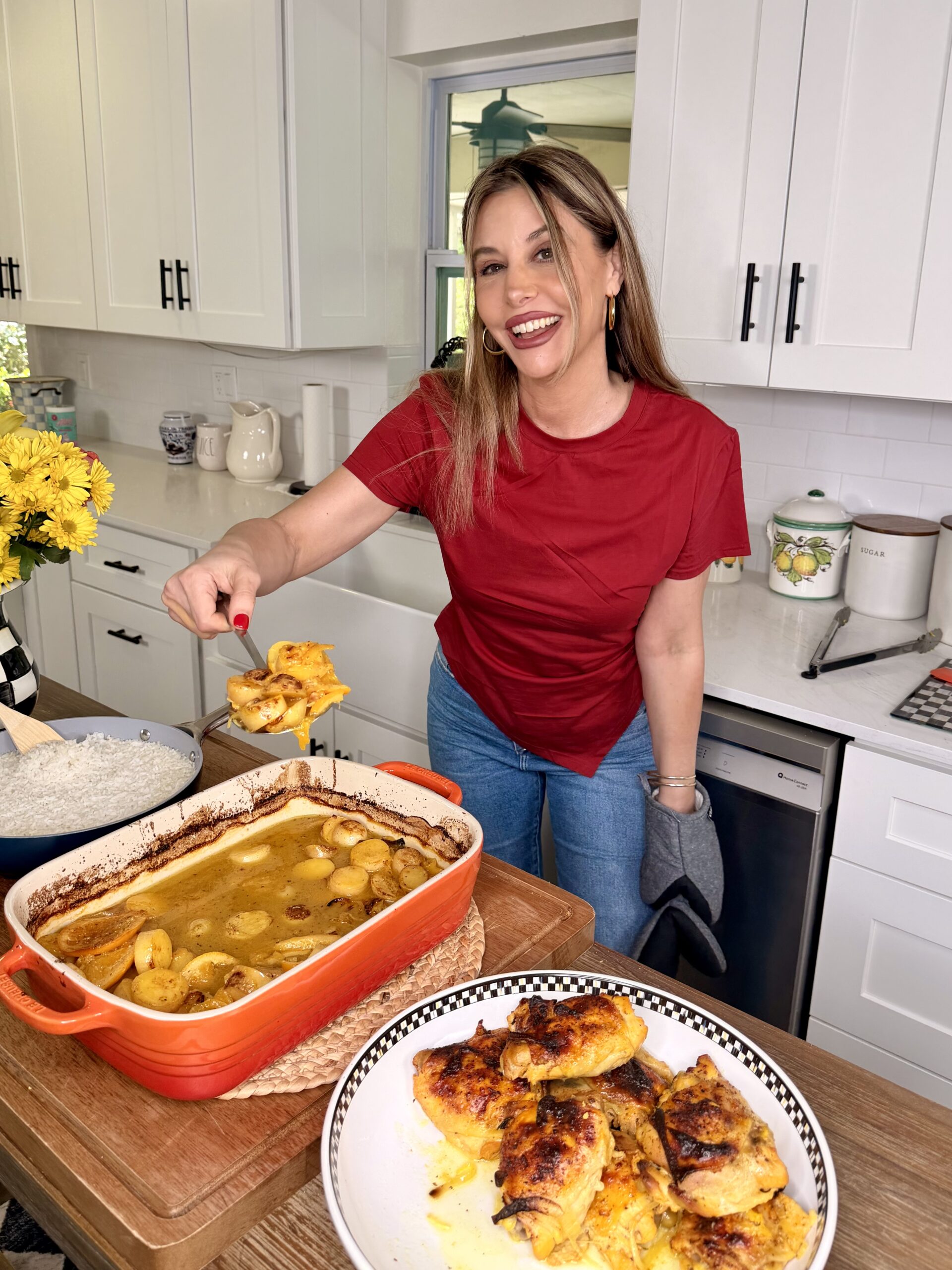 A woman in a red shirt smiles while serving cheesy casserole from a baking dish in a bright kitchen. Next to her, a plate of One-Pan Mojo Chicken & Potatoes sits on the counter, ready to enjoy.