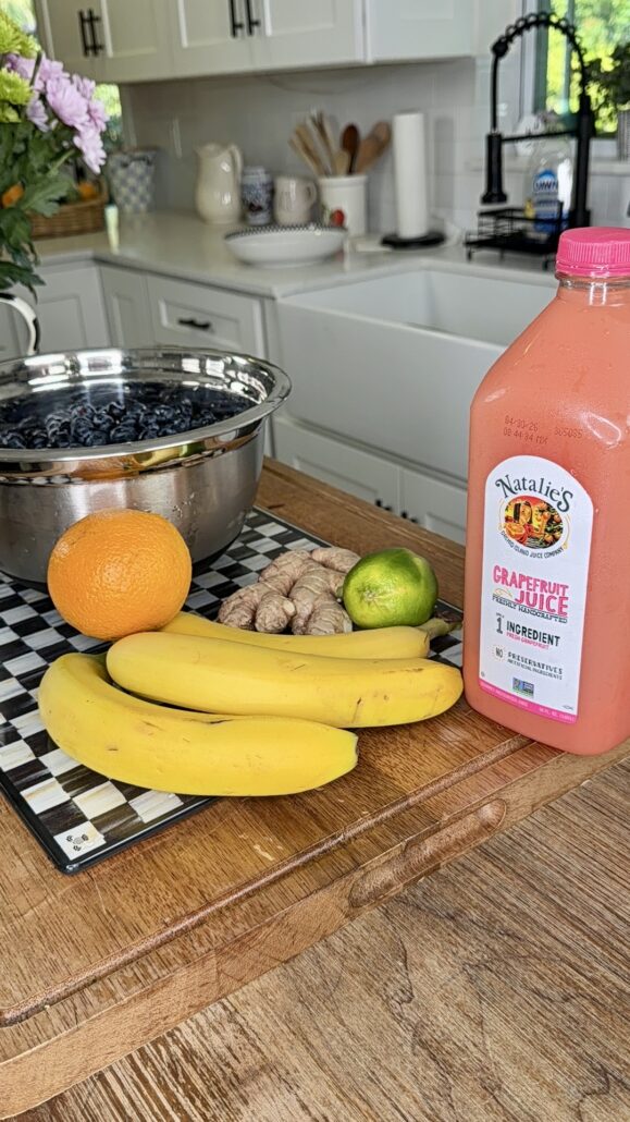 A kitchen counter with bananas, an orange, a lime, ginger root, a large bowl of blueberries, and a bottle of Natalie’s grapefruit juice. White cabinets and kitchenware are visible in the background.