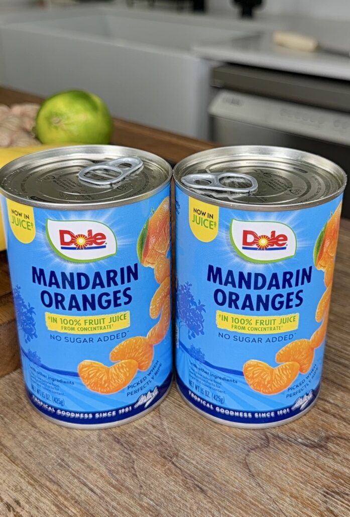 Two cans of Dole Mandarin Oranges in juice, with blue labels and images of mandarin slices, are placed side by side on a wooden surface. A lime and part of a lemon are visible in the background.