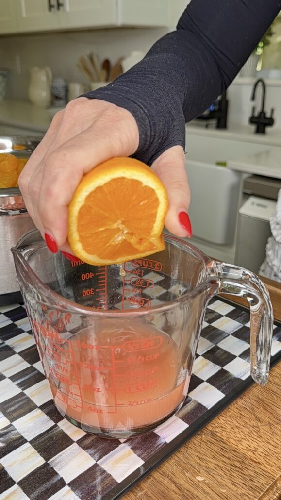 A person with red nail polish squeezes half an orange over a glass measuring cup on a checkered cloth, collecting fresh juice on a wooden countertop in a kitchen.