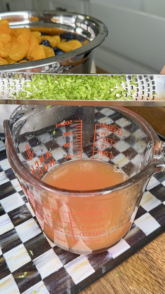 A metal grater with green zest rests over a glass measuring cup containing pink juice. In the background, a bowl of sliced mandarin oranges and blueberries is visible. The scene is set on a checkered surface.