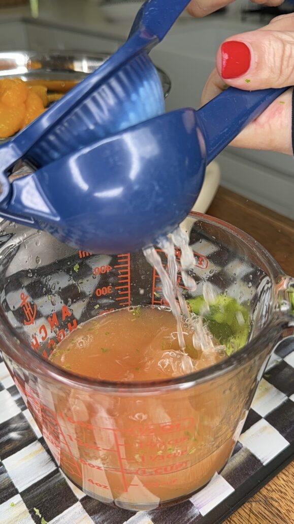A person with red nail polish squeezes a blue citrus juicer over a glass measuring cup filled with juice, on a checkered cloth.