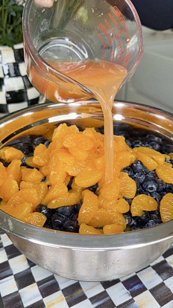 A glass measuring cup pours orange juice over a stainless steel bowl filled with blueberries and mandarin orange segments on a checkered surface.