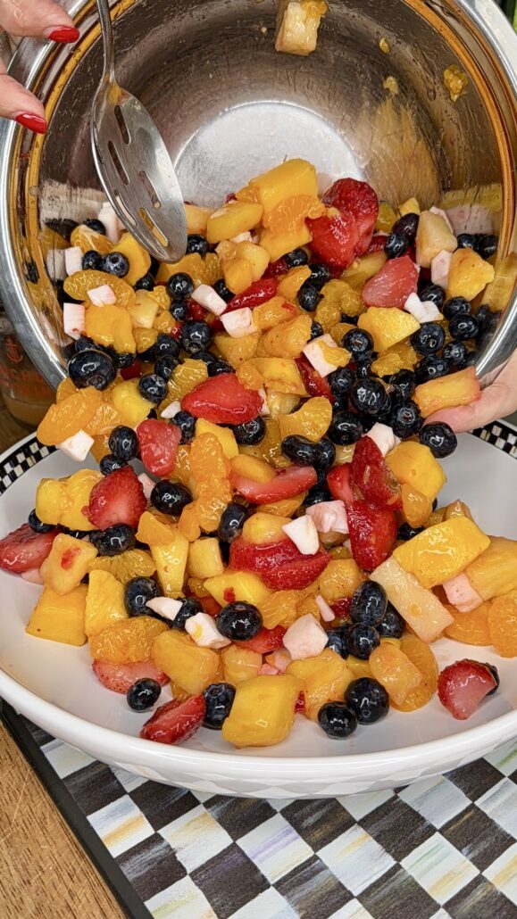 A close-up of a person pouring a colorful fruit salad with strawberries, blueberries, peaches, and small white cubes from a metal bowl into a white serving dish.