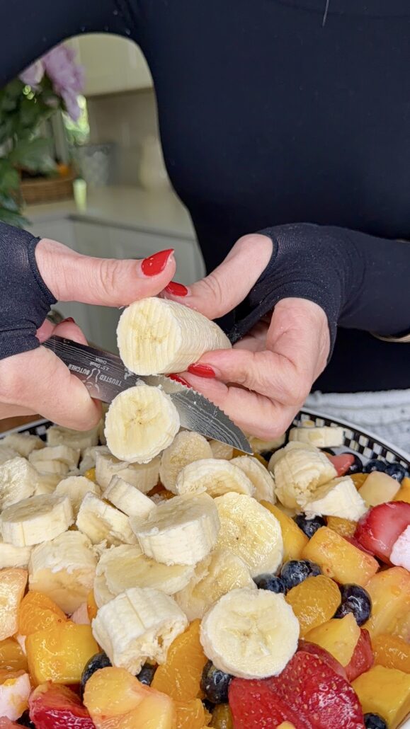 A person wearing black gloves is slicing a banana over a bowl of mixed fruit, including strawberries, blueberries, peaches, and more banana slices. The persons nails are painted red.