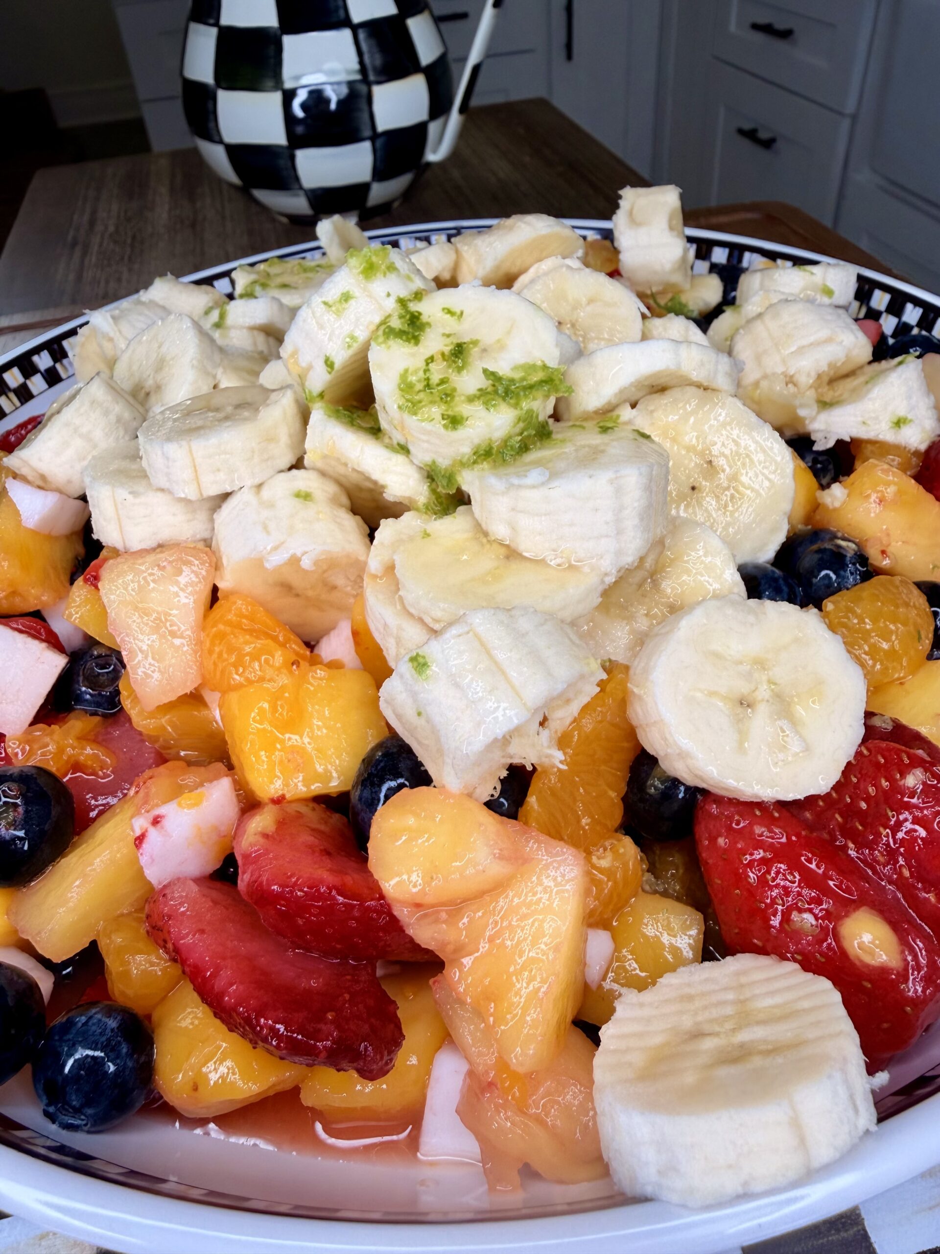 A close-up of an Easy Tropical Fruit Salad featuring sliced bananas, strawberries, blueberries, peaches, and melon in a bowl, topped with grated lime zest. A checkered teapot is in the background.