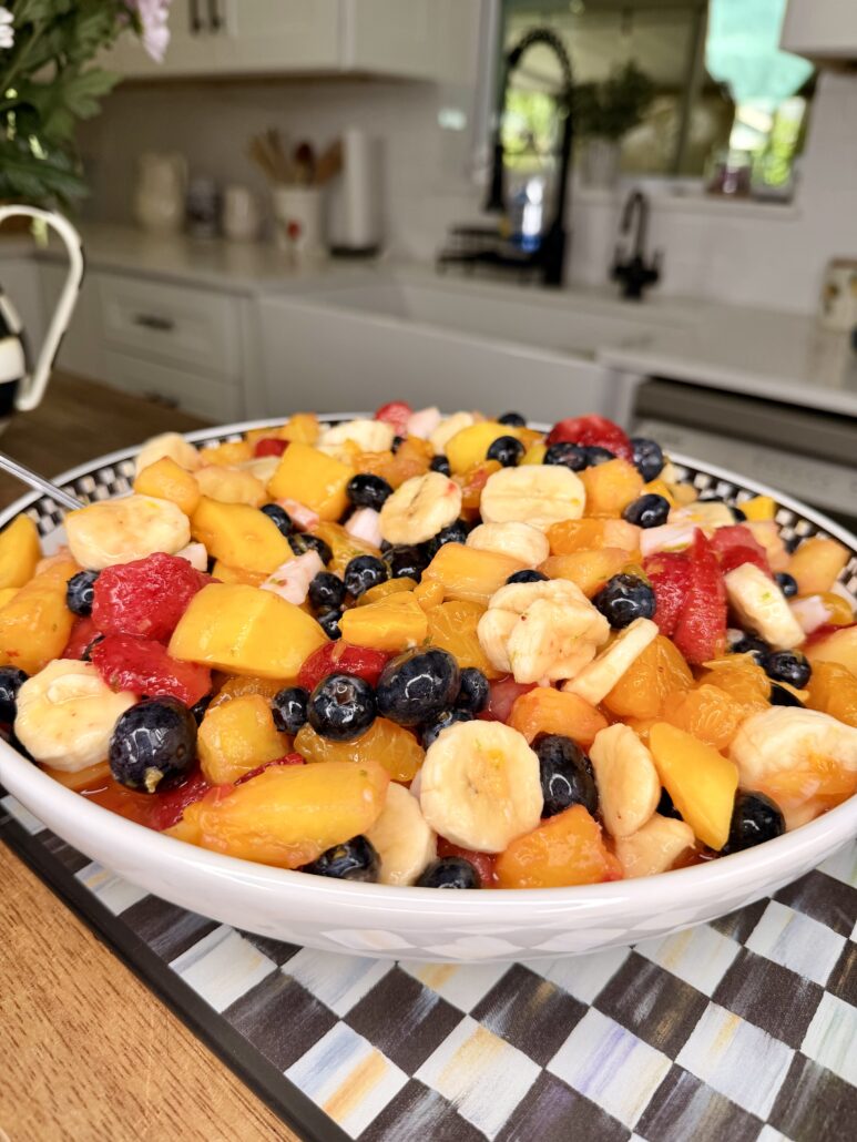 A large bowl of colorful fruit salad with sliced bananas, peaches, strawberries, blueberries, and grapes sits on a striped placemat in a bright kitchen.