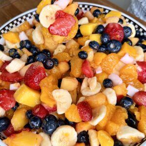 A close-up of an Easy Tropical Fruit Salad featuring bananas, strawberries, blueberries, mandarin oranges, and mango pieces in a bowl. A fork is lifting a portion of the vibrant mixed fruit above the bowl.