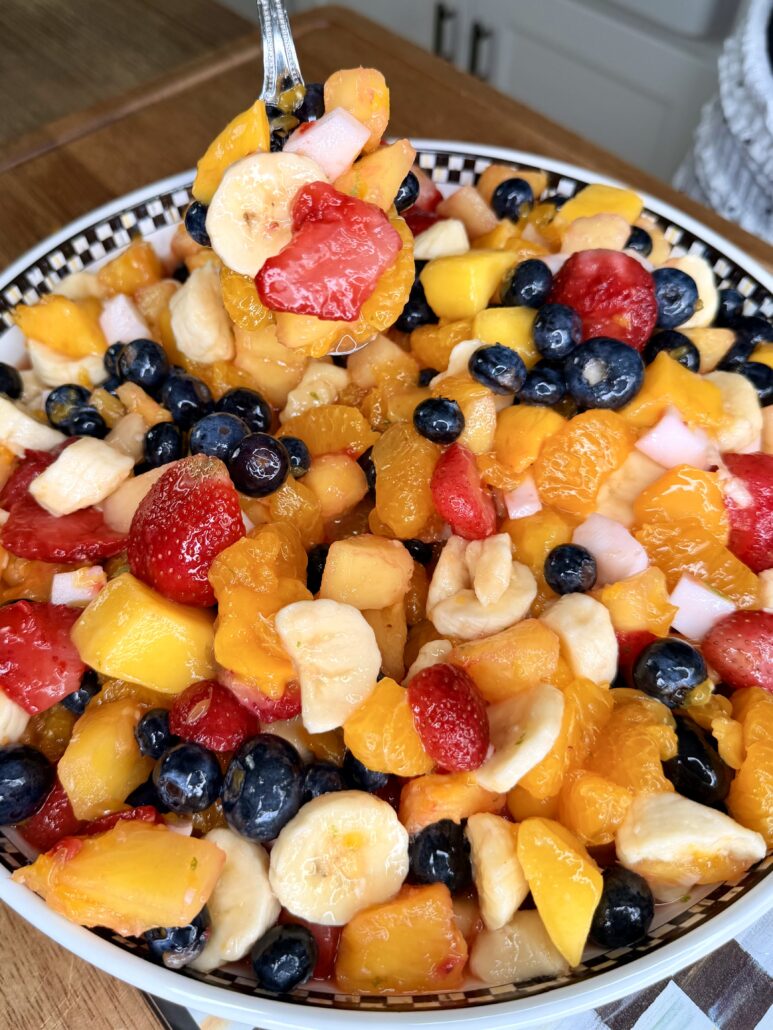 A colorful fruit salad in a bowl, featuring sliced bananas, strawberries, blueberries, mangoes, oranges, and melon. A fork is lifting a serving from the bowl. The salad sits on a wooden table.