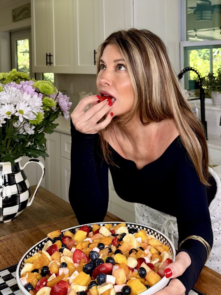 A woman with long hair and red nails is eating a strawberry in a kitchen. She holds a spoon over a large bowl of colorful fruit salad resting on a counter next to a vase of flowers.