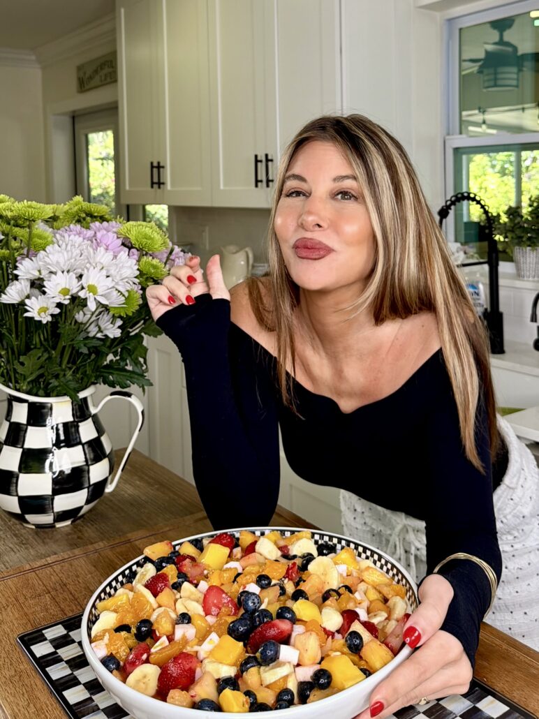 A smiling woman with long hair and red nails leans on a kitchen counter, holding a spoon. In front of her is a large bowl of colorful fruit salad. A vase of flowers and a checkered pitcher sit nearby.